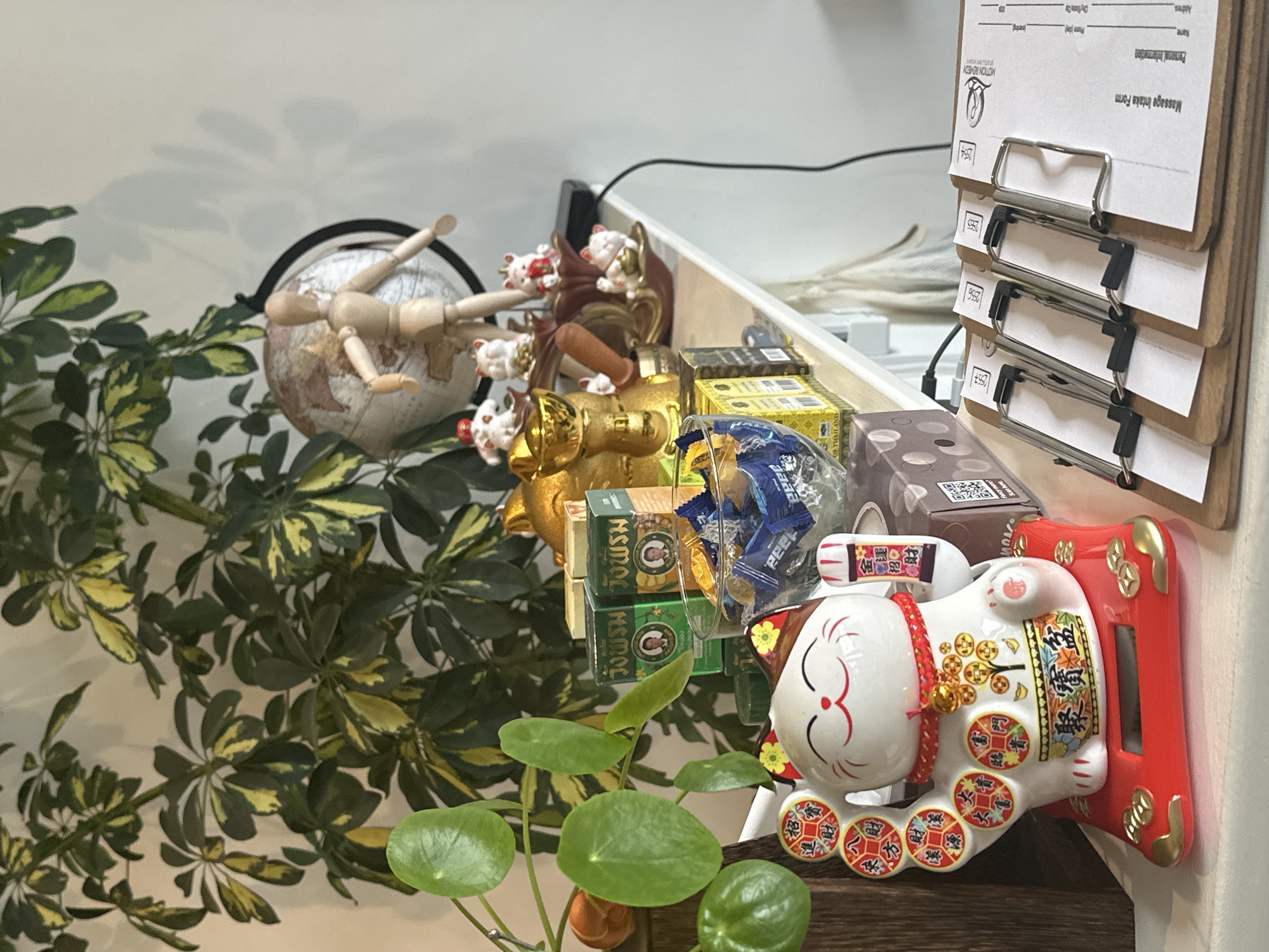 The image depicts an interior scene, specifically a counter or shelf, within a commercial establishment identified as "MOTION REMEDY" by text on forms. This business is "Motion Remedy Thai Massage," located in London, United Kingdom.
In the foreground, a white ceramic Maneki-neko (waving cat) figurine, adorned with red and gold accents, is positioned on a red solar-powered base. Its left paw is raised, displaying eight circular gold coins with Chinese characters, and its right paw holds a small red and gold scroll. To the right of the figurine, a stack of four wooden clipboards is present. Each clipboard holds a white "Massage Intake Form" with the "MOTION REMEDY" logo—a stylized figure in a healing pose—and is numbered sequentially (e.g., "253", "254", "255", "256"). A brown cardboard box with white polka dots and a visible QR code is behind these clipboards.
On a higher shelf in the background, against a plain white wall, a variety of items are arranged. These include a golden Buddha statue, several smaller white Maneki-neko figurines with red accents, and a clear glass bowl filled with individually wrapped candies (blue and yellow wrappers). Multiple green and gold boxes, identified by Thai script as "วังพรม" (Wang Prom, a Thai herbal balm brand), are also on display. Further back on the shelf, a small globe is situated behind a wooden artist's mannequin. A variegated green and yellow leafy plant (likely Schefflera arboricola 'Variegata') and a green Pilea peperomioides plant are visible on the left side of the frame. A black power cable descends from a device on the right side of the shelf. No individuals are present in the scene.