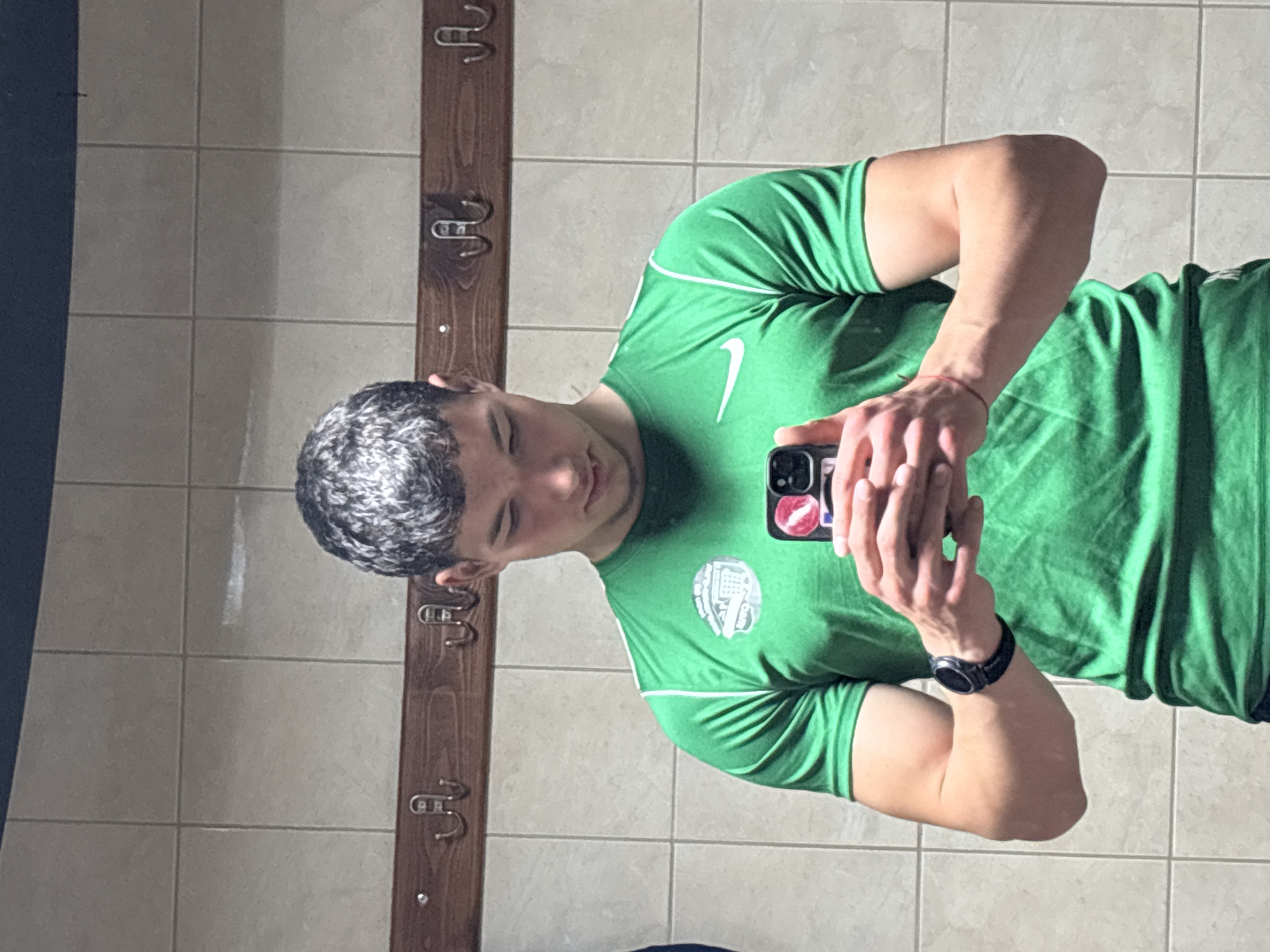 A person stands in a tiled room, possibly a bathroom or locker room, taking a selfie in a mirror. The person is wearing a green athletic t-shirt with white trim and a white Nike logo. The person is also wearing a black watch on their left wrist and is holding a black smartphone. The background includes a tiled wall and a wooden rack with hooks. The location cannot be verified as being in Zlatograd, Bulgaria from the image alone.