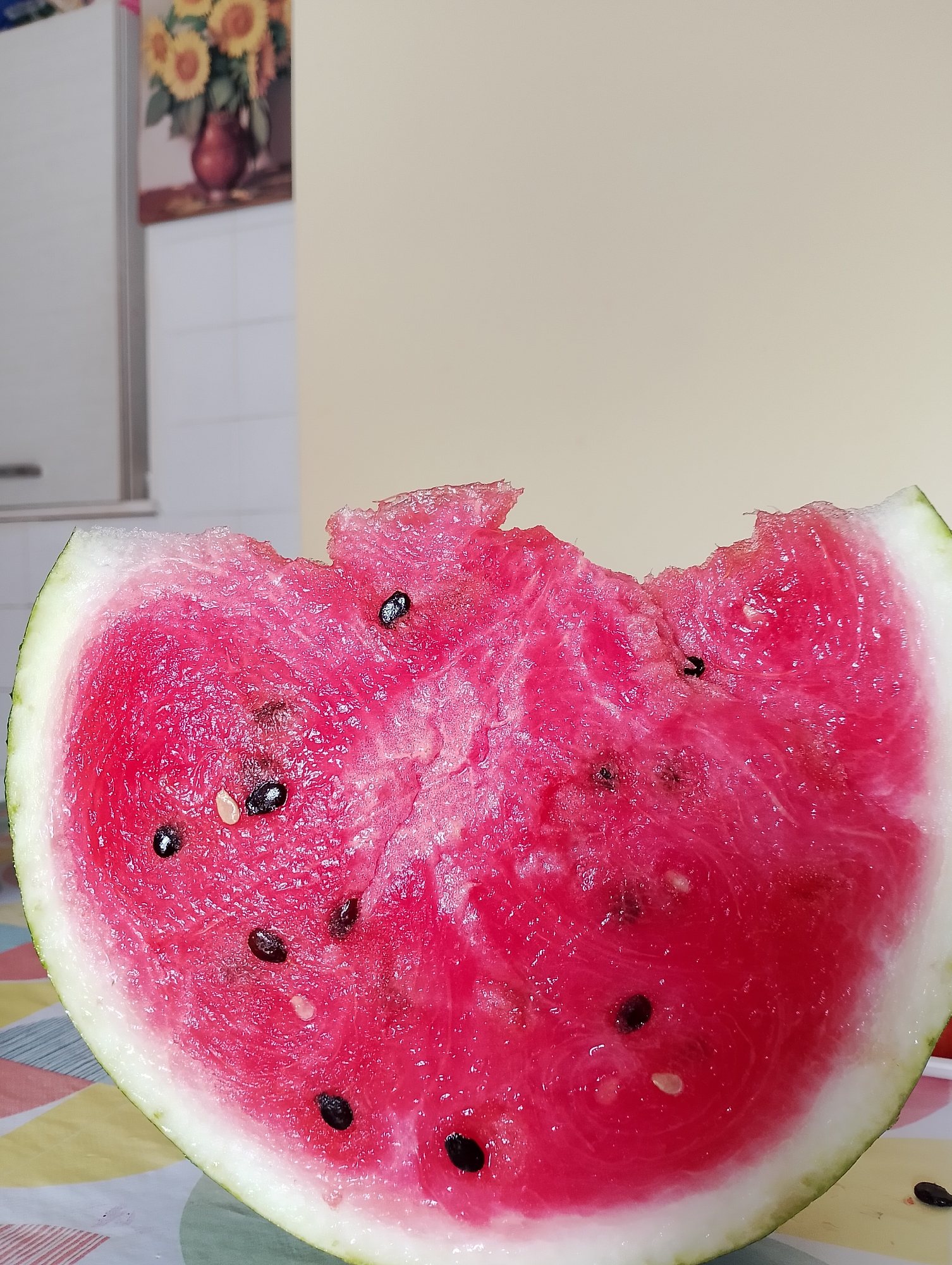 A close-up shot focuses on a slice of watermelon on a table covered with a colorful, patterned tablecloth. The watermelon slice is a vibrant pink, with several black seeds visible. The top edge of the watermelon appears to have been bitten or broken off. In the background, a painting of sunflowers in a vase is hanging on a wall, with a white tiled surface below it, possibly a backsplash. The overall environment suggests a domestic setting, likely a kitchen or dining area. The location is not specified in the image or the provided information.