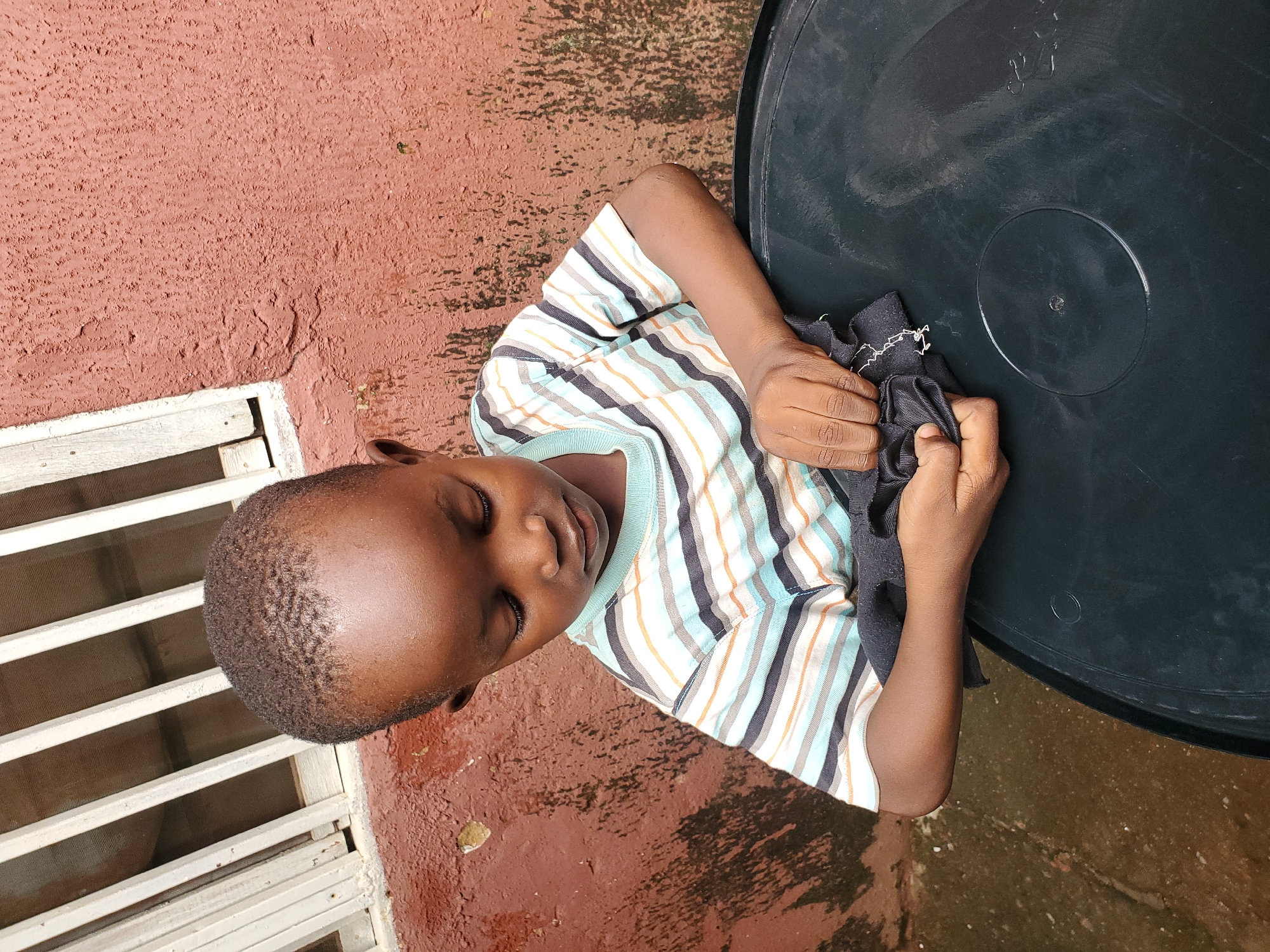 A young Black boy with short, dark hair and closed eyes rests his head on his arm, which is positioned on a large, dark circular object. He is wearing a striped t-shirt with horizontal bands of white, light blue, beige, and dark gray. His right hand is holding a crumpled piece of black fabric. The boy is lying on a reddish-brown, textured surface that appears to be concrete or stucco. To his left, a portion of a white metal grate or window frame is visible. The ground is wet in places, suggesting recent rain. The setting appears to be outdoors, possibly a patio or courtyard, with the reddish surface and the dark circular object suggesting an outdoor appliance or furniture. The lighting and the wet ground suggest it might be a humid day. The boy's relaxed posture and closed eyes convey a sense of peace or rest. There is no visible text in the image.