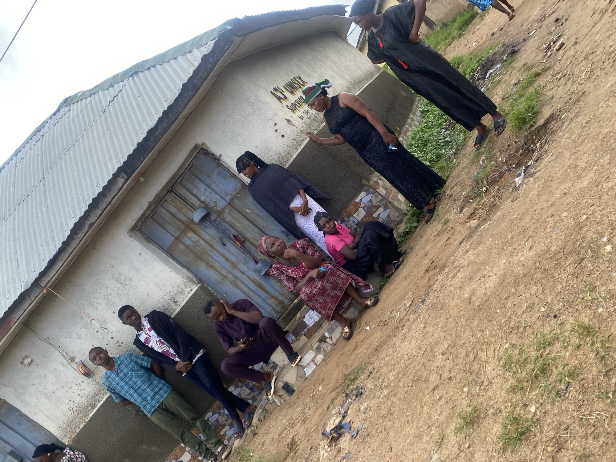 In the city of Jos, Nigeria, a group of nine individuals are gathered outside a building with a corrugated metal roof. The building appears to be a small shop or business, with the name "AD UNISEX" visible on the wall. The ground in front of the building is dirt and gravel, with some patches of grass.
The group consists of five men and four women, all dressed in casual clothing. Two young men in the foreground are leaning against the wall, one wearing a blue plaid shirt and the other a black jacket over a patterned shirt. Behind them, a man in a maroon outfit is sitting and looking at his phone. To the right, a woman in a red and white patterned dress is seated, looking to the side. Another woman in a pink shirt and black pants is crouching nearby. Further to the right, a woman in a black dress with a colorful headscarf is gesturing with her hand. Two other women in black traditional attire are also present, one standing and the other partially visible. The overall atmosphere appears relaxed, with the individuals engaged in conversation or simply resting.