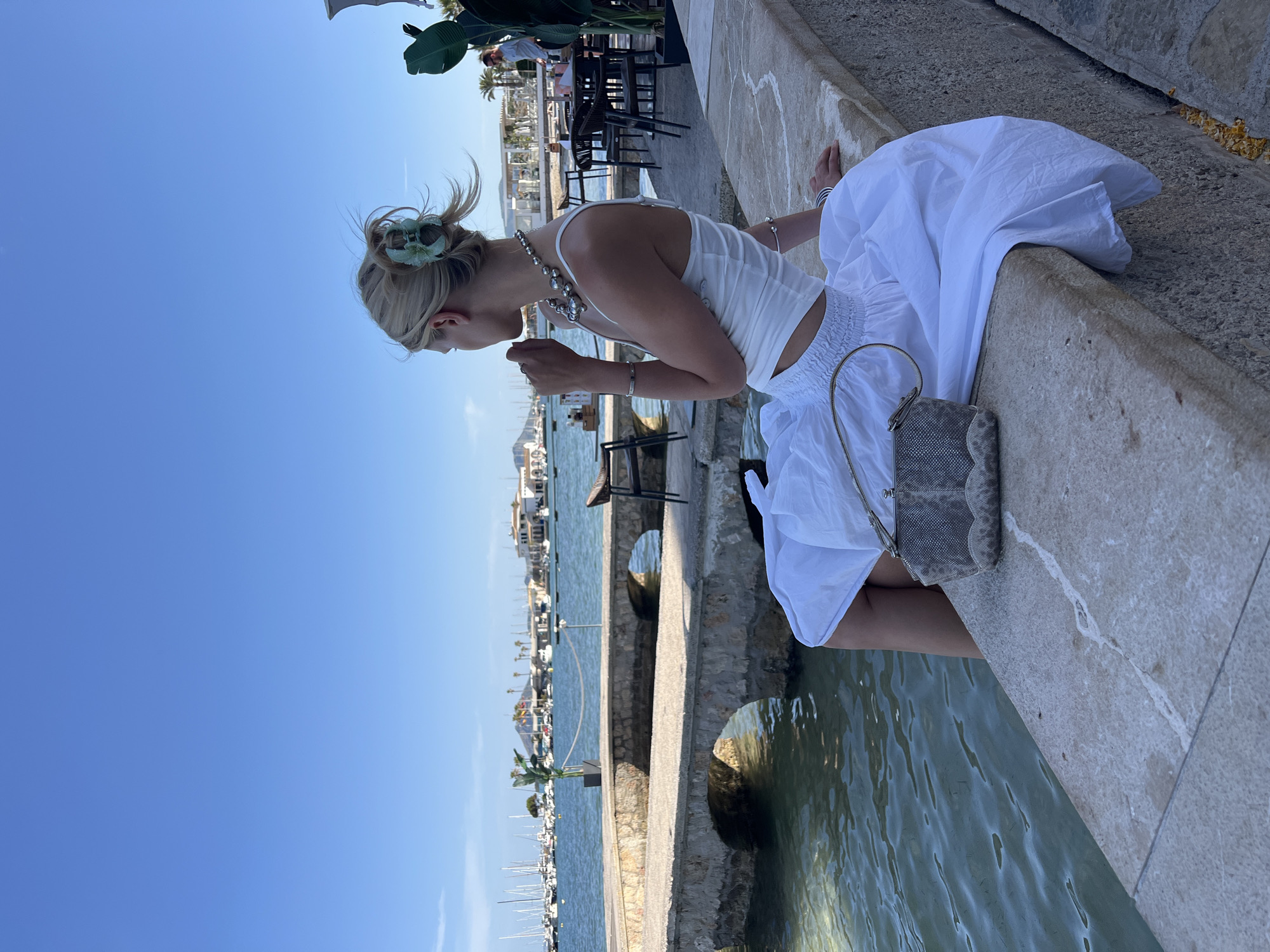 A young woman with blonde hair pulled up in a bun, secured by a light green claw clip, sits on a stone ledge overlooking a body of water in the city of Pollença, Spain. She is wearing a white tank top and a long, flowing white skirt. A silver beaded necklace adorns her neck, and she wears a silver bracelet on her left wrist. In her lap rests a small, textured grey handbag. The water is calm and clear, with small waves lapping against the stone structure. In the background, a harbor filled with sailboats is visible, with buildings and palm trees lining the shore. A stone bridge with two archways spans a section of the water in the mid-ground. Several outdoor tables and chairs are set up on a paved area to the right of the woman. The sky is a clear, bright blue.