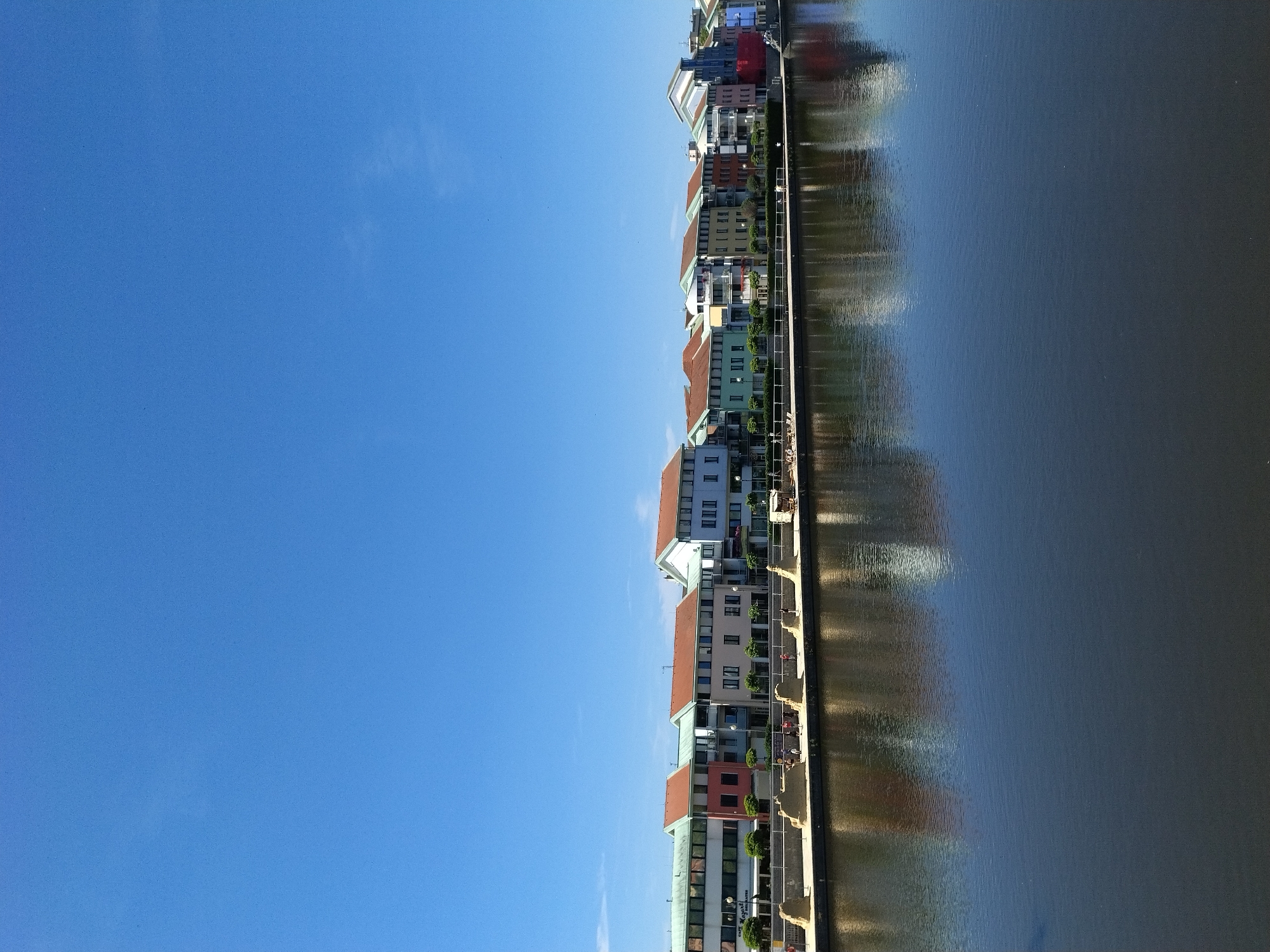 The image depicts a sunny day in Písek, Czechia, showcasing a waterfront with modern buildings reflecting in the dark water. A clear, bright blue sky dominates the upper portion of the frame. Along the waterfront, a row of multi-colored, multi-story buildings is visible, featuring a variety of architectural styles and roof designs. These buildings appear to be residential or commercial properties. In front of the buildings, a paved walkway runs along the water's edge, lined with trees and decorative sculptures or structures. Several people can be seen walking and standing on the walkway. The water is calm, with clear reflections of the buildings and the sky. The overall scene suggests a vibrant urban environment with a focus on waterfront development.
