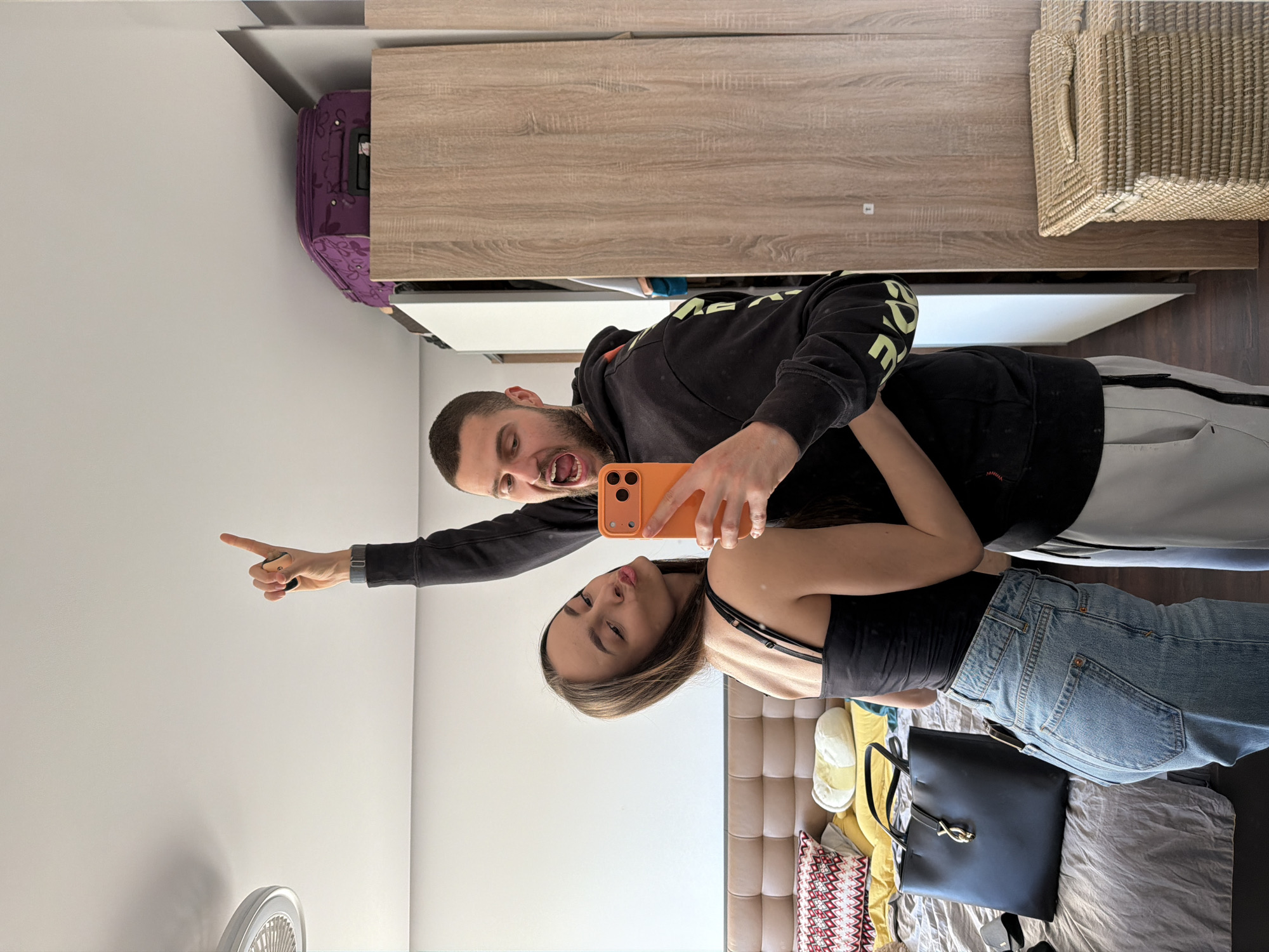 A man and a woman are taking a selfie in a room. The room contains a light-colored wooden cabinet, a purple suitcase, a wicker basket, a bed with a tufted headboard, a black handbag, clothing, and a fan. The man is wearing a black hoodie and light-colored pants, and is pointing to the upper left. The woman is wearing a black top and blue jeans. The location appears to be a bedroom in a residential building in Sofia, Bulgaria.