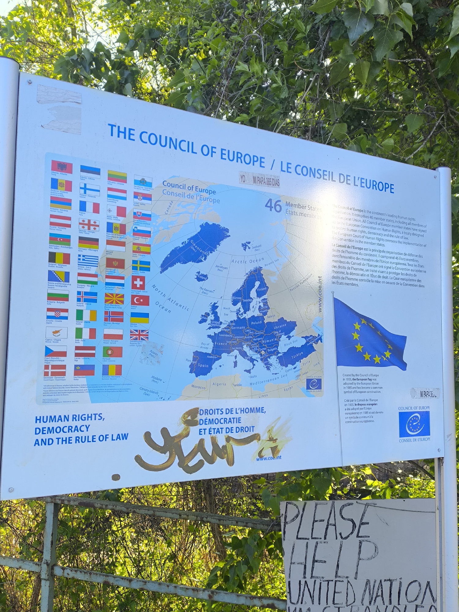The image shows a sign for The Council of Europe located outdoors, likely in Strasbourg, France, the seat of the Council. The sign features the flags of the member states, a map of Europe, and information about the Council in English and French. The sign is vandalized with graffiti in gold paint. Below the Council of Europe sign, there's another sign with the handwritten words "PLEASE HELP UNITED NATION" followed by illegible words. The signs are situated in front of some greenery, suggesting a park or garden setting. The sign structure is metallic, showing signs of rust.