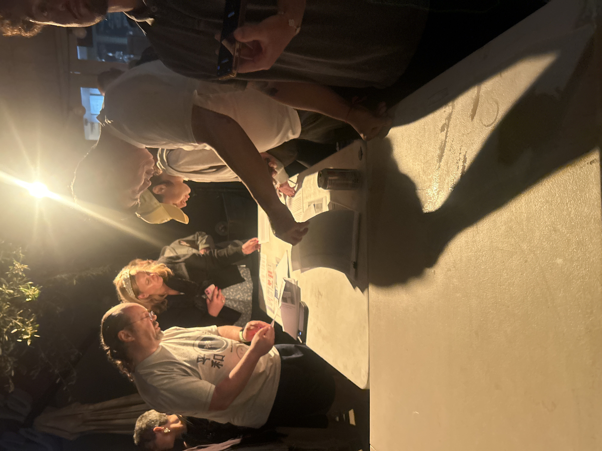 A group of people are gathered around a table outdoors, possibly at night, given the lighting. The table appears to be covered with papers and documents, and the people seem to be examining them closely. One person is holding a cell phone, possibly taking pictures of the documents. Another individual is wearing a t-shirt with Japanese characters. The scene is lit by an external light source. There is no immediate indication of the exact location within Los Angeles.