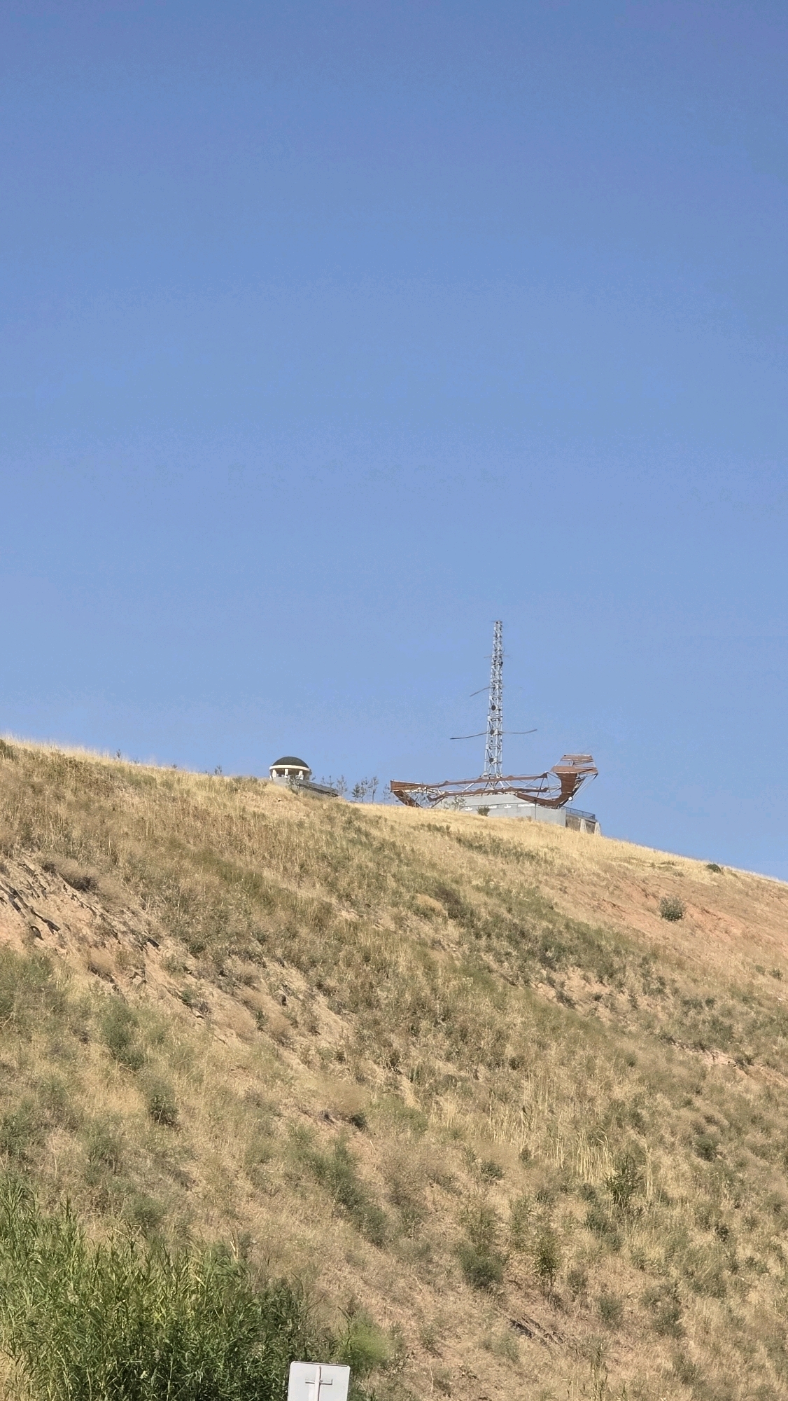 The image shows a wide shot of a dry, grassy hillside under a clear blue sky. On the crest of the hill, two structures are visible. To the left, a small, domed building with white walls is situated. To the right of this building, a tall, metallic structure resembling a ship's mast and hull stands. The ship-like structure appears to be made of metal beams and has an incomplete or skeletal appearance. The hillside is covered in dry, yellow-brown grass and sparse green shrubs. The overall environment suggests a semi-arid or arid climate. The location is identified as Алатау ауылдық округі, Kazakhstan. There are no people or animals visible in the image.
