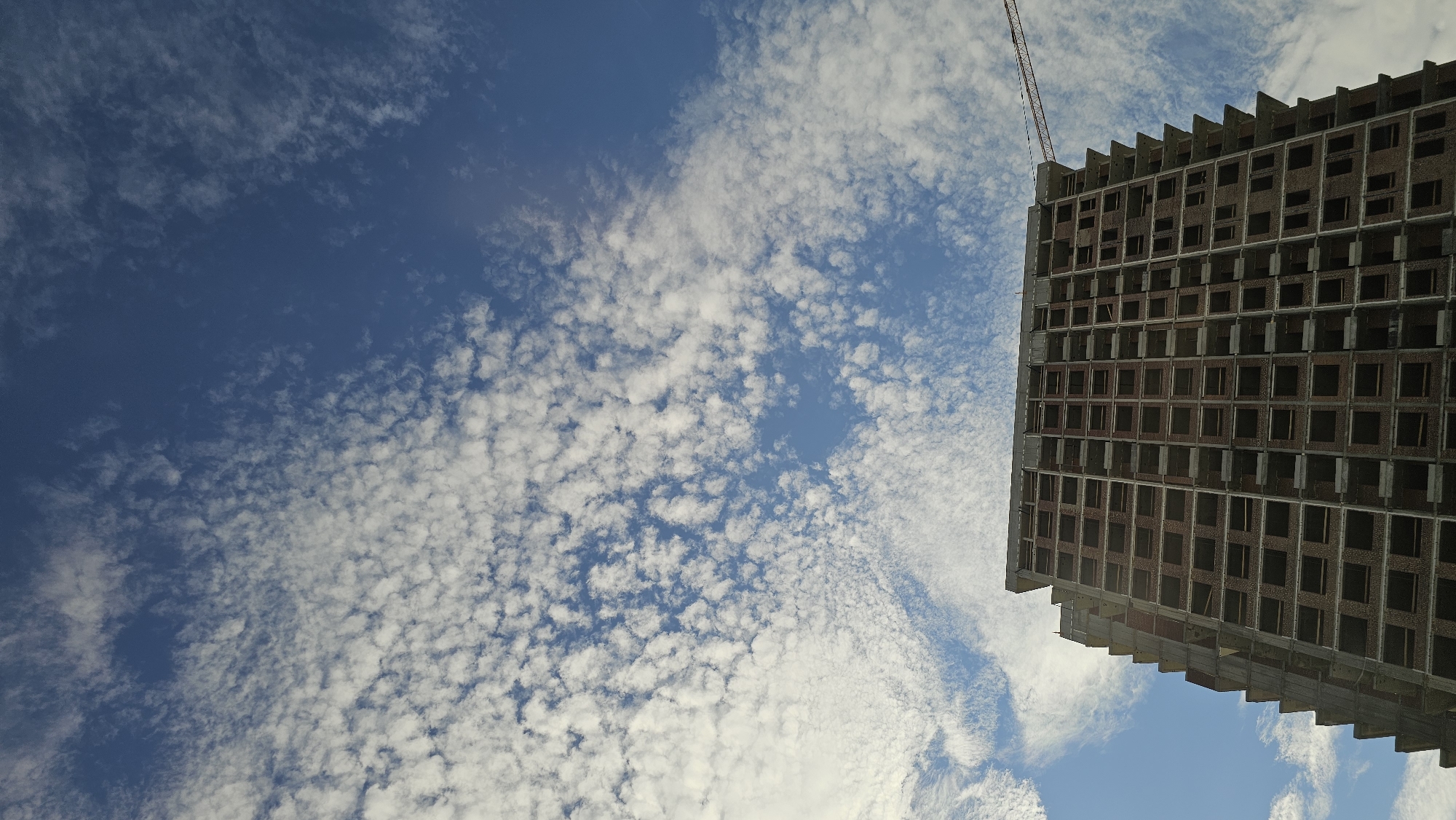 A low-angle shot captures the stark facade of an unfinished skyscraper against a bright blue sky filled with scattered, wispy clouds. The building's raw concrete structure is visible, with rows upon rows of dark, empty window frames creating a repeating grid pattern. The top of the building features jagged, unfinished edges, hinting at its incomplete state. A tall construction crane extends from the upper right corner of the frame, its thin boom reaching towards the sky, signifying ongoing construction. The clouds are spread across the sky, with some appearing as small, puffy masses and others as elongated streaks. The lighting suggests it is daytime, likely with clear weather. The image composition emphasizes the scale of the building and its contrast with the natural expanse of the sky. No people are visible in the frame. The scene is likely a construction site in Yekaterinburg, Russia, given the provided location context.