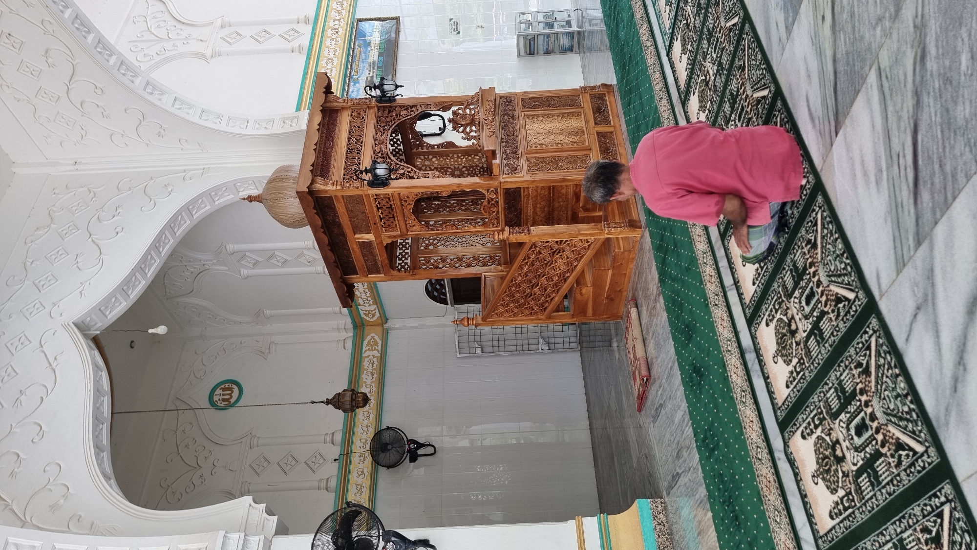 This is a photograph taken from a low angle inside a mosque in Lapang Timu, Indonesia. The mosque features white, ornate walls with Arabic calligraphy and decorative arches. In the center of the frame is a tall, intricately carved wooden minbar (pulpit). A man wearing a pink shirt and a checkered sarong is sitting on a prayer mat on the green carpeted floor, facing the minbar. There are fans mounted on the walls and a light fixture hanging from the ceiling. A small prayer mat is placed on the floor in front of the minbar. The floor is tiled with light-colored marble. The overall atmosphere suggests a place of worship.