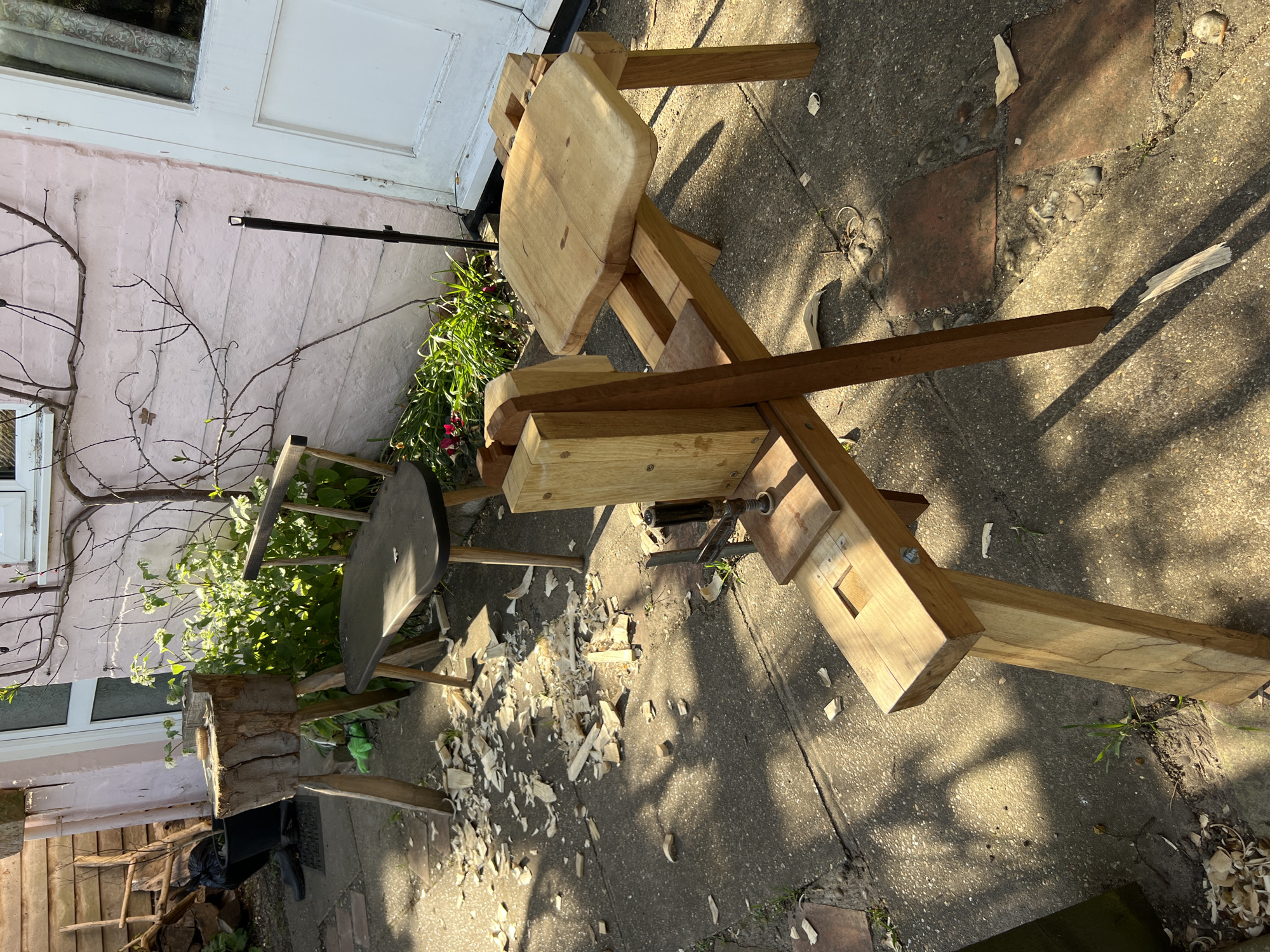 The image shows an outdoor woodworking setup in East Suffolk, United Kingdom. A shave horse and stump stand are present on a concrete surface, scattered with wood shavings. A pink brick building with a white door is visible in the background, along with vegetation. There are no people visible in the image.
