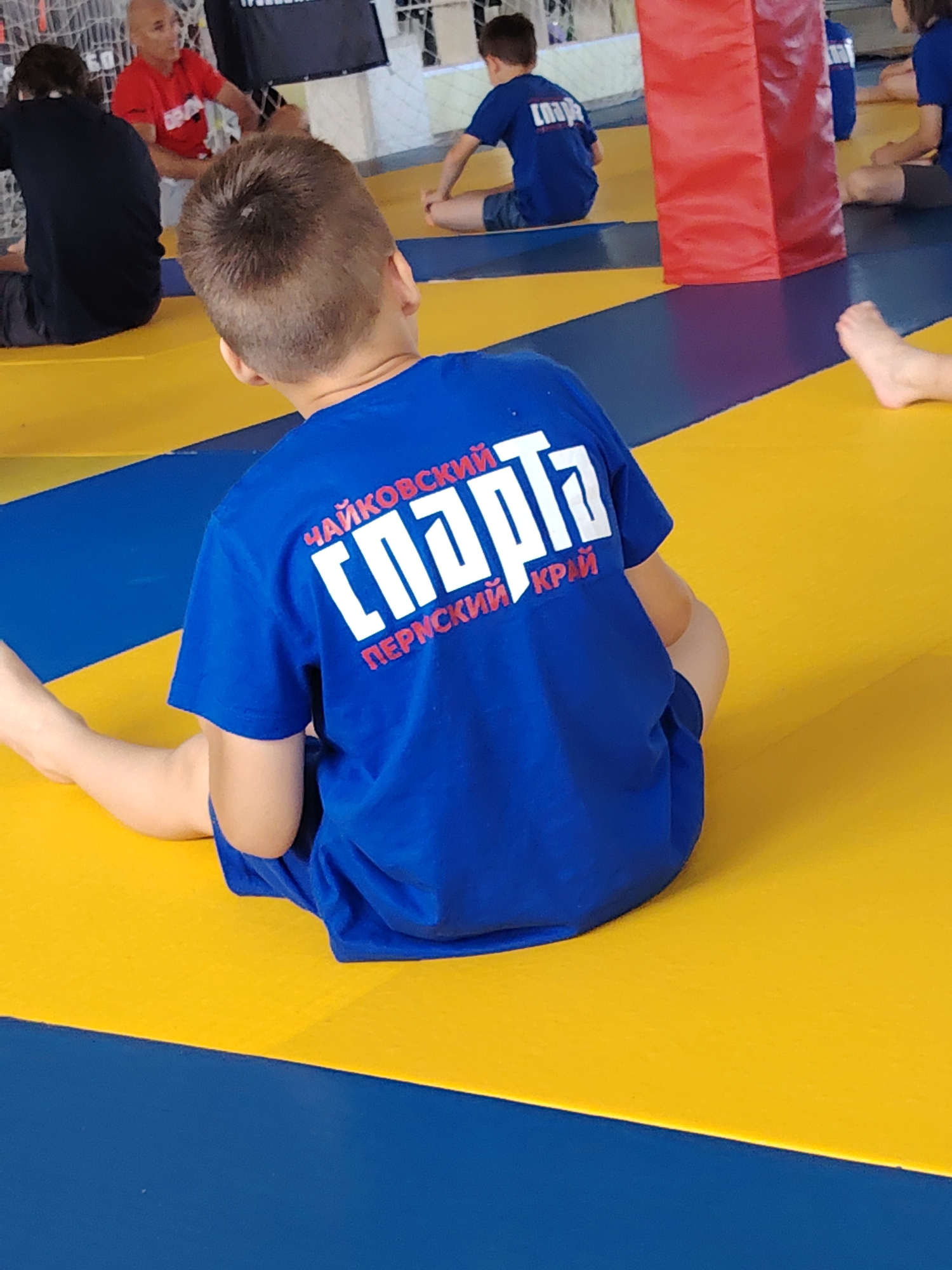 The image shows a group of young people sitting on a mat, likely during a sports or training session. The mat is yellow and blue. A boy in the foreground is wearing a blue t-shirt with white lettering. The lettering appears to be in Russian and says "ЧАЙКОВСКИЙ СПАРТА ПЕРМСКИЙ КРАЙ," which translates to "Chaykovsky Spartak Perm Krai." This suggests the location is a sports facility in Chaykovsky, Perm Krai, Russia, associated with the Spartak sports club. Other individuals are visible in the background, also appearing to be participants in a similar activity. One adult in a red shirt is also present. There is a red padded pillar or wall in the mid-ground. The setting appears to be an indoor sports venue.
