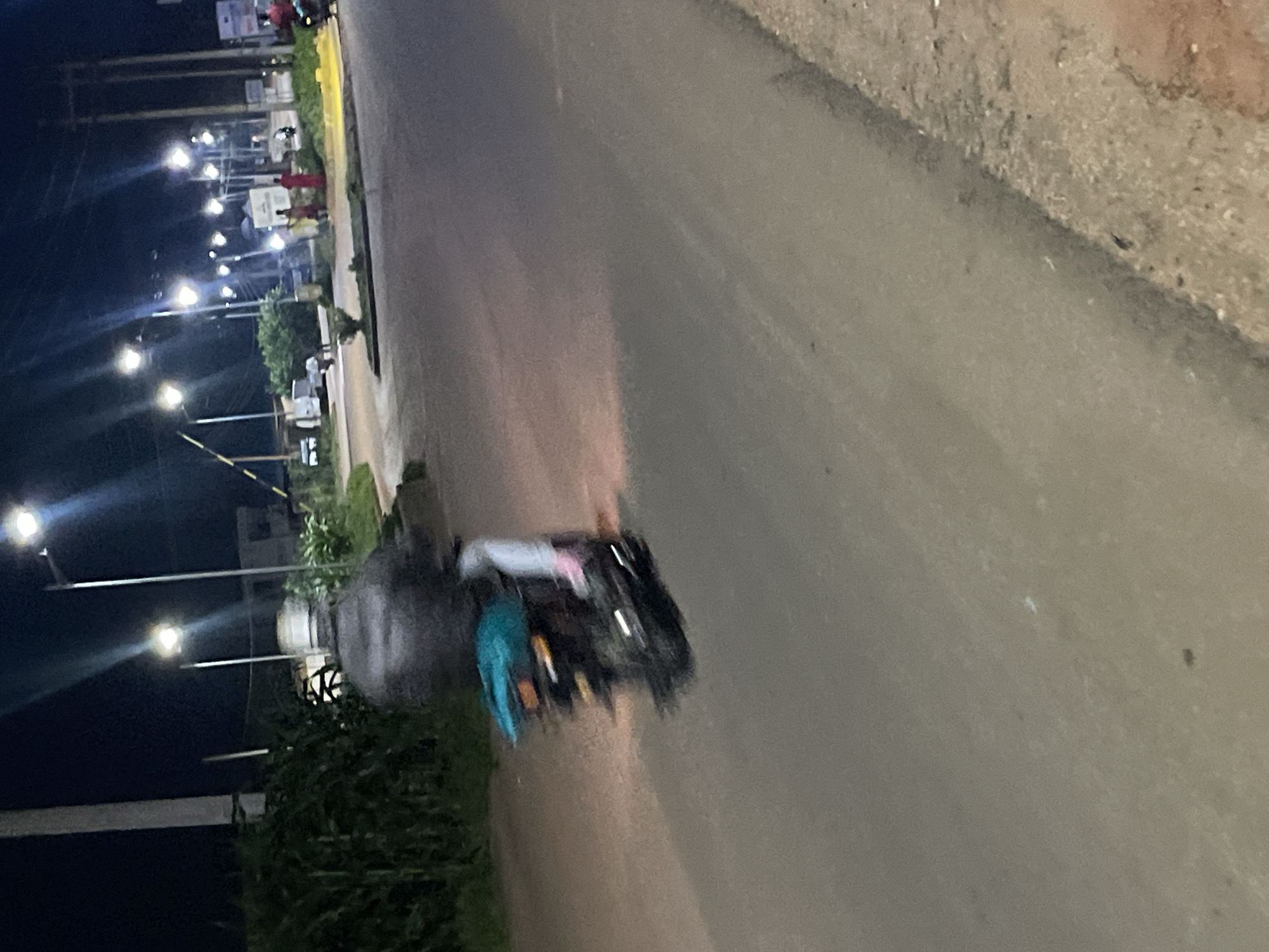 A medium shot shows a motorcycle with a rider from the side as it travels down a paved road at night. The motorcycle has a light blue tarp or bag secured to the back. The rider is wearing a dark jacket and a helmet. The road is dimly lit by streetlights and vehicle headlights, and there is some vegetation visible on the left side of the image. The scene appears to be in an urban or suburban area. The location is Barakin Lerawi, Nigeria.