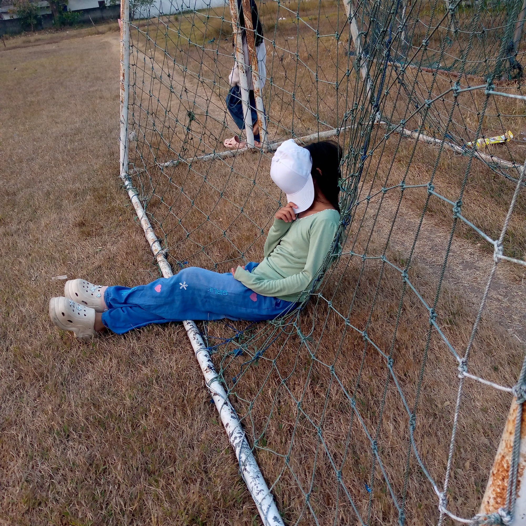 In Pacing, Indonesia, a young girl wearing a light green long-sleeved shirt, blue pants with a pink heart and a white flower embroidered on them, and white clogs rests on the ground leaning against a green soccer net. She is wearing a white baseball cap, obscuring her face. Her legs are extended, and her feet are positioned towards the left side of the frame. Another person, partially visible and wearing blue jeans and pink sandals, stands behind her. The scene takes place on a dry, grassy field, likely a soccer field, with a weathered white pole supporting the net. The lighting suggests it is late afternoon or early evening.