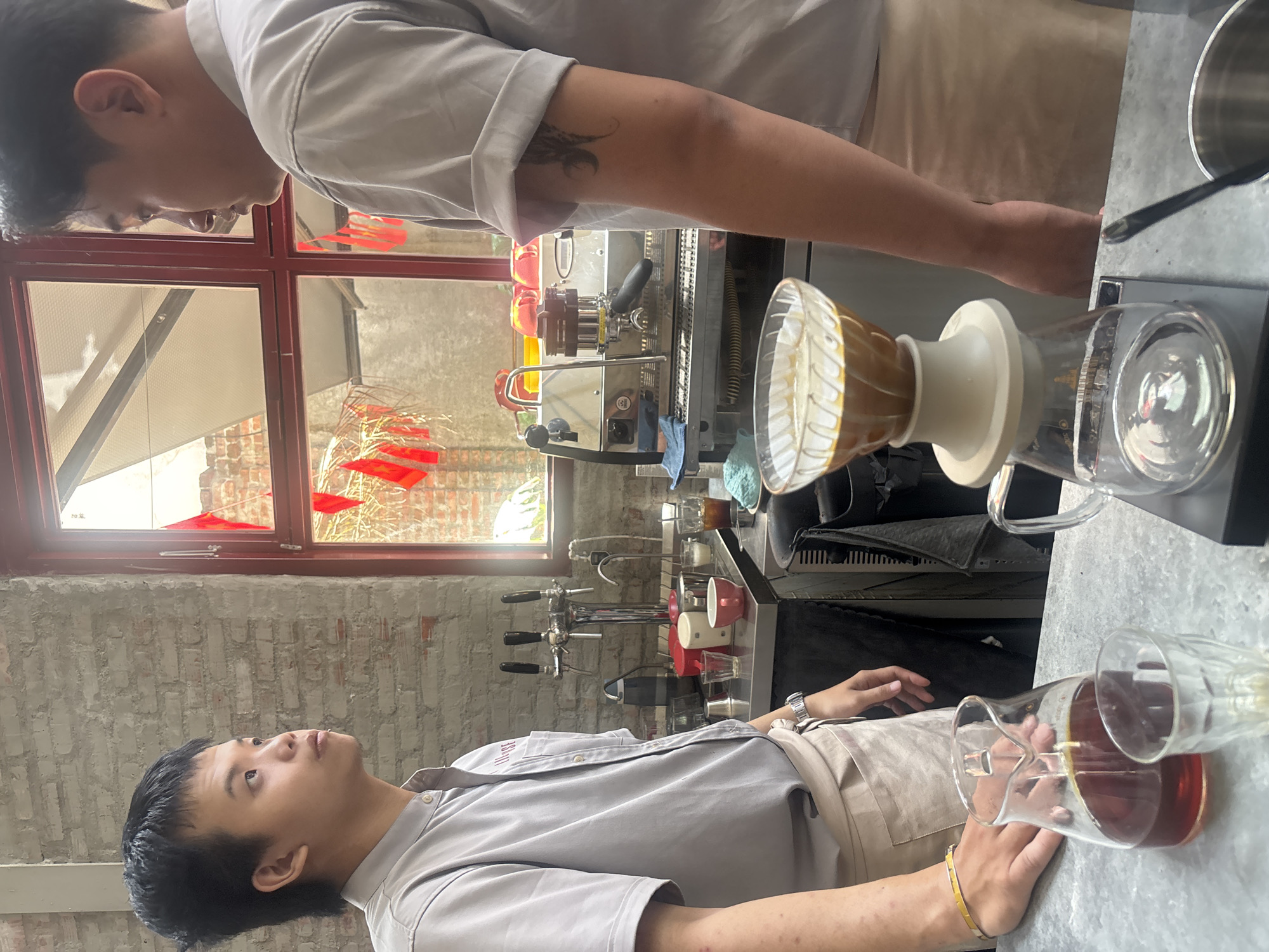 The image depicts two individuals, likely baristas, working behind a counter in a coffee shop located in Đà Nẵng, Vietnam. The shop has an industrial aesthetic, with exposed brick walls and red-trimmed windows. One barista, on the left, is wearing a light gray shirt and a beige apron, with black hair and a focused expression as he looks off to the side. He has a yellow bracelet on his left wrist. The other barista, on the right, is partially visible, also wearing a light gray shirt and beige apron, with a tattoo on his right arm. He appears to be in conversation with the first barista.
In the foreground, a pour-over coffee setup is visible, with a glass dripper filled with coffee and a carafe below. A separate glass carafe containing brewed coffee and a small glass are also on the counter. Behind the baristas, a professional espresso machine is present, along with various coffee-making equipment, including taps for draft beverages, cups, and small pitchers. Red flags, possibly national or decorative, are hanging outside the window. The overall atmosphere suggests a bustling coffee preparation area.
