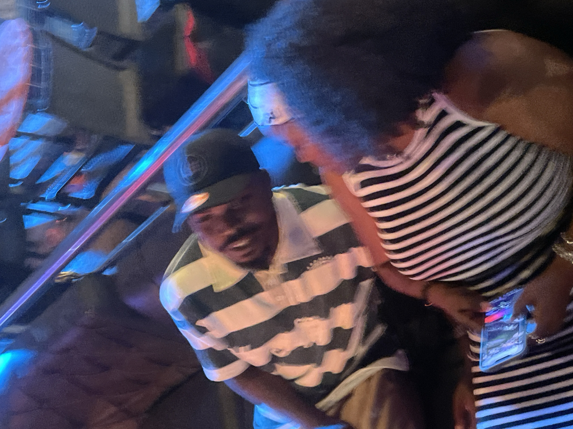Two individuals, one with dark skin and wearing a black and white striped shirt and a black baseball cap, appears to be looking down and to the right, possibly in conversation. The other individual, with dark skin and curly dark hair, is wearing a black and white striped shirt and is holding a smartphone. They are seated in what appears to be an indoor setting with blurred background elements that suggest neon lighting and seating structures, possibly a bar or club. The overall impression is a candid moment captured in a dimly lit, vibrant environment. The location is specified as Guzape, Nigeria.