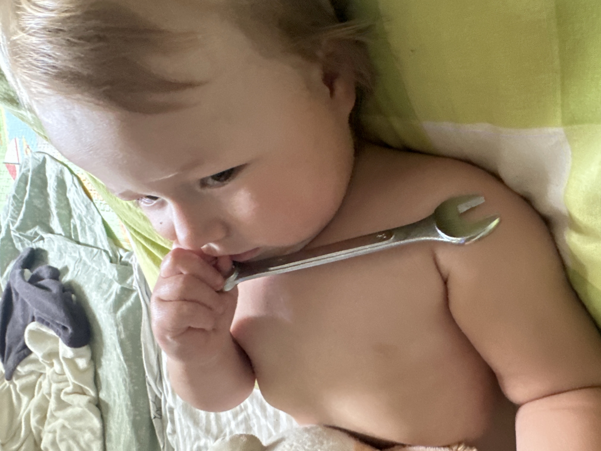 This close-up, overhead shot features a baby lying down, with a metal wrench resting on their upper body. The baby, with fair hair, is looking downwards and towards the left, with their right hand near their mouth, appearing to be touching it with the wrench. The baby is unclothed from the chest down. The background consists of bedding with a light green and white checkered pattern, with some crumpled fabric visible to the left, including a dark gray garment. The lighting suggests it might be daytime, possibly indoors. No visible text is present in the image. The overall impression is of a candid moment of a baby playing with an unexpected object.