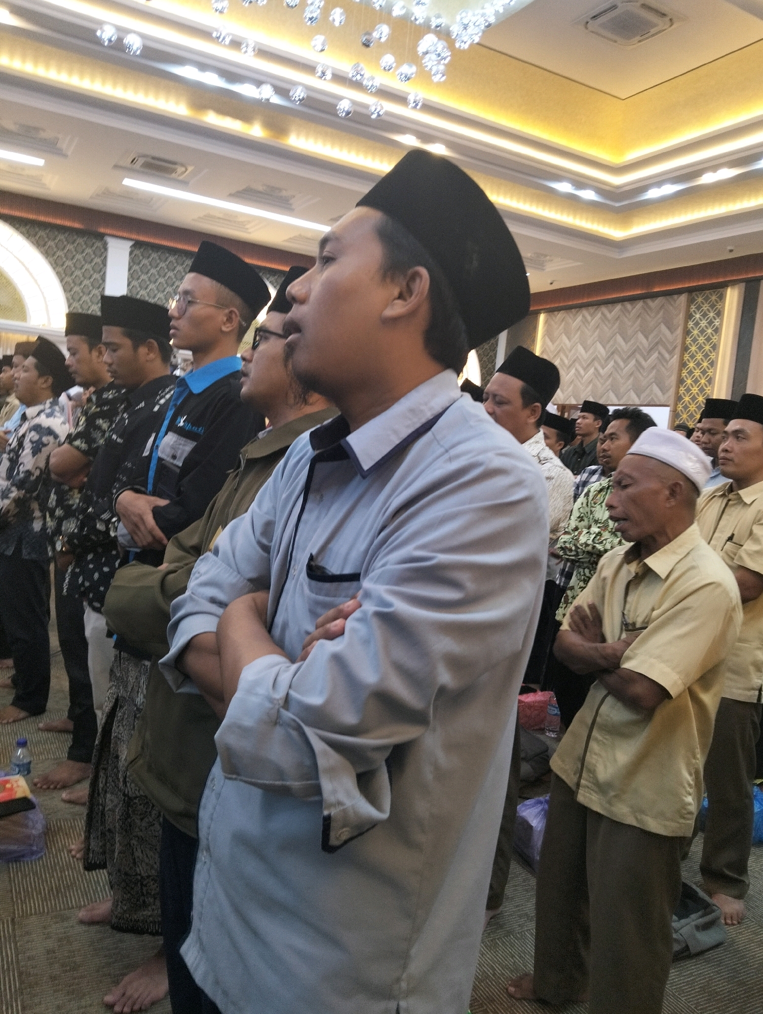 The image shows a group of men standing in a hall, possibly for a religious gathering. They are all wearing traditional attire, including peci (caps) and long-sleeved shirts. Some are wearing sarongs. The men are standing in rows, facing the same direction, with their arms folded. The hall appears to be well-lit, with modern ceiling lights and decorative elements. The floor is carpeted. In the foreground, a man in a light blue shirt is prominently featured, with his arms folded. In the background, more men are visible, some with eyeglasses, and a ceiling fan. The overall atmosphere suggests a formal or ceremonial event. The image does not contain any explicit visual cues that would pinpoint the exact location within Paciran, Indonesia, other than it is an indoor setting.