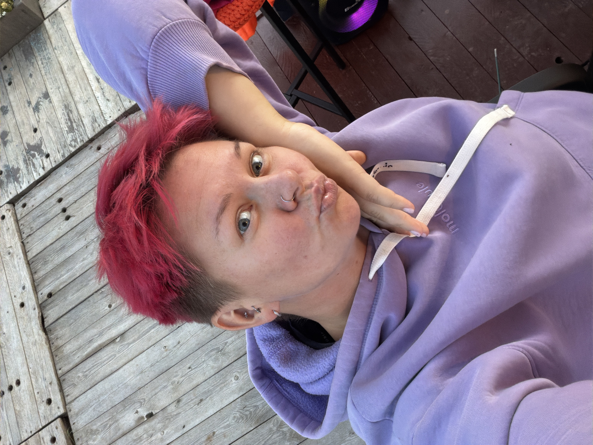 A woman with short, vibrant pink hair is lying down on a wooden deck, resting her head on her arm. She is wearing a light purple hoodie with the drawstrings visible. Her lips are puckered, and she is looking towards the camera with blue eyes. She has piercings on her ear and nose. The background shows a wooden outdoor surface and a dark chair. A portable speaker with purple and pink lights is visible on the right side of the image. The image appears to have been taken outdoors during daylight.