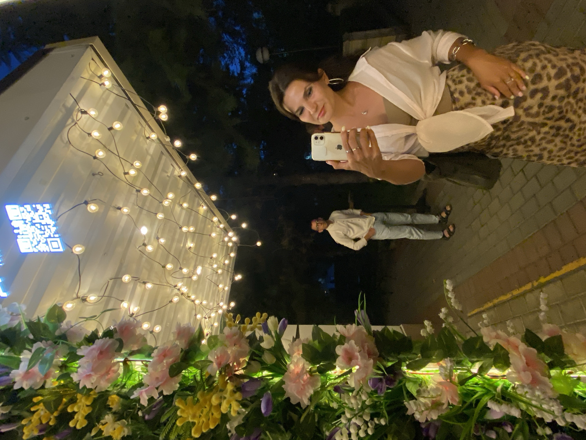 A woman is taking a selfie with a smartphone in her right hand, holding it up and slightly to her left. She is looking at the phone with a slight smile and her head tilted. She is wearing a white open shirt tied at the waist over a light brown tank top, a leopard print skirt, and several rings and bracelets. Her left hand is resting on her stomach. A dark bag is slung over her left shoulder. Behind her, to the left, a man wearing a light-colored button-down shirt and light-colored pants stands with his hands on his hips. He is facing the same direction as the woman but looking away from the camera. A large floral arrangement, featuring pink, yellow, and white flowers, is positioned on the left side of the frame. The scene appears to be outdoors at night, with string lights illuminating a white ceiling or overhang above the subjects. The ground is paved with brick-like stones. The background is dark and appears to be foliage. There is a QR code visible on the white ceiling. The location is stated to be Feodosia, Ukraine.