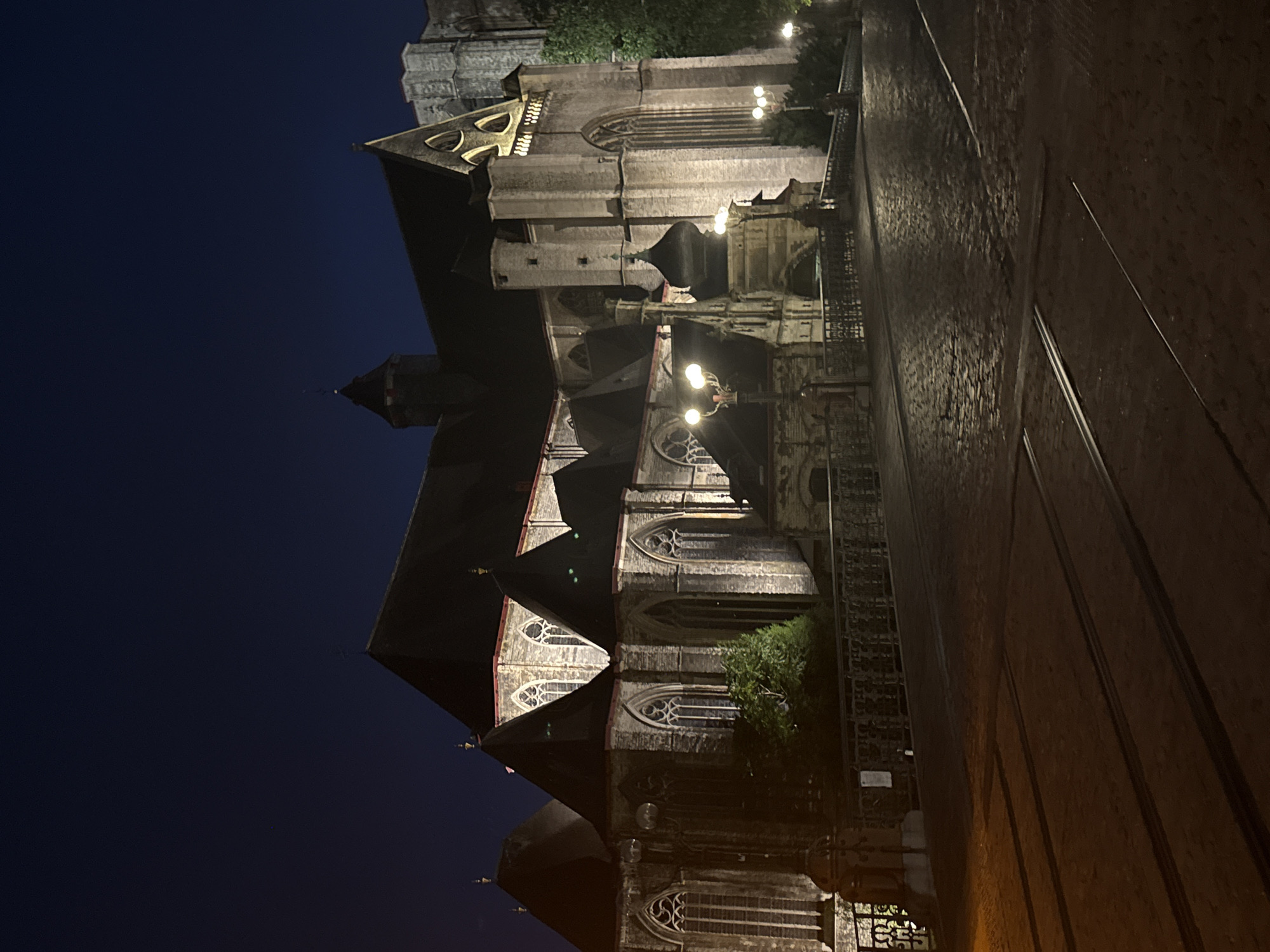 The image depicts a nighttime view of the St. Bavo's Cathedral in Ghent, Belgium. The cathedral is a large Gothic structure, illuminated by artificial lighting that highlights its stone facade and architectural details, including pointed arches, stained glass windows, and steep gabled roofs. A cobblestone street with tram tracks runs in the foreground, leading towards a bridge that crosses a canal or body of water, partially visible on the left. The bridge is adorned with decorative metal railings and lampposts that cast pools of light onto the street. Trees and other foliage are visible around the cathedral and along the canal. The sky is dark, indicating nighttime. There are no people visible in the image.