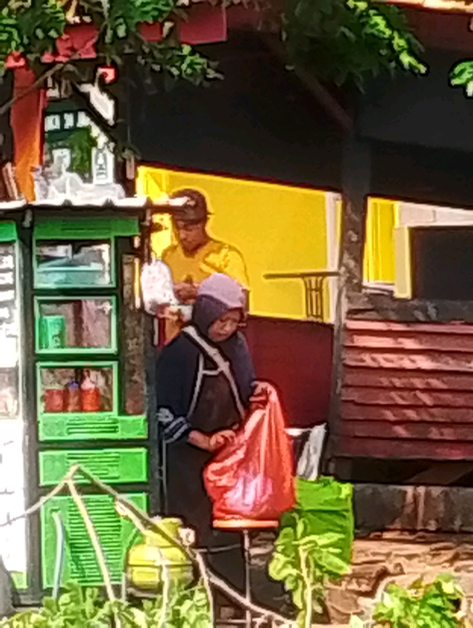 This image depicts a street food stall in Probolinggo, Indonesia. Two individuals are present. A man wearing a dark cap and a yellow t-shirt stands behind the stall. A woman in a purple head covering and dark apron is in front of the stall, holding a bright orange plastic bag. The stall is green with multiple shelves displaying various items. A yellow gas cylinder is visible to the left of the stall. The background shows a building with yellow and maroon walls, and a dark roof. Lush green plants are in the foreground, suggesting an outdoor setting. The scene appears to be a typical day at a local market or roadside vendor.