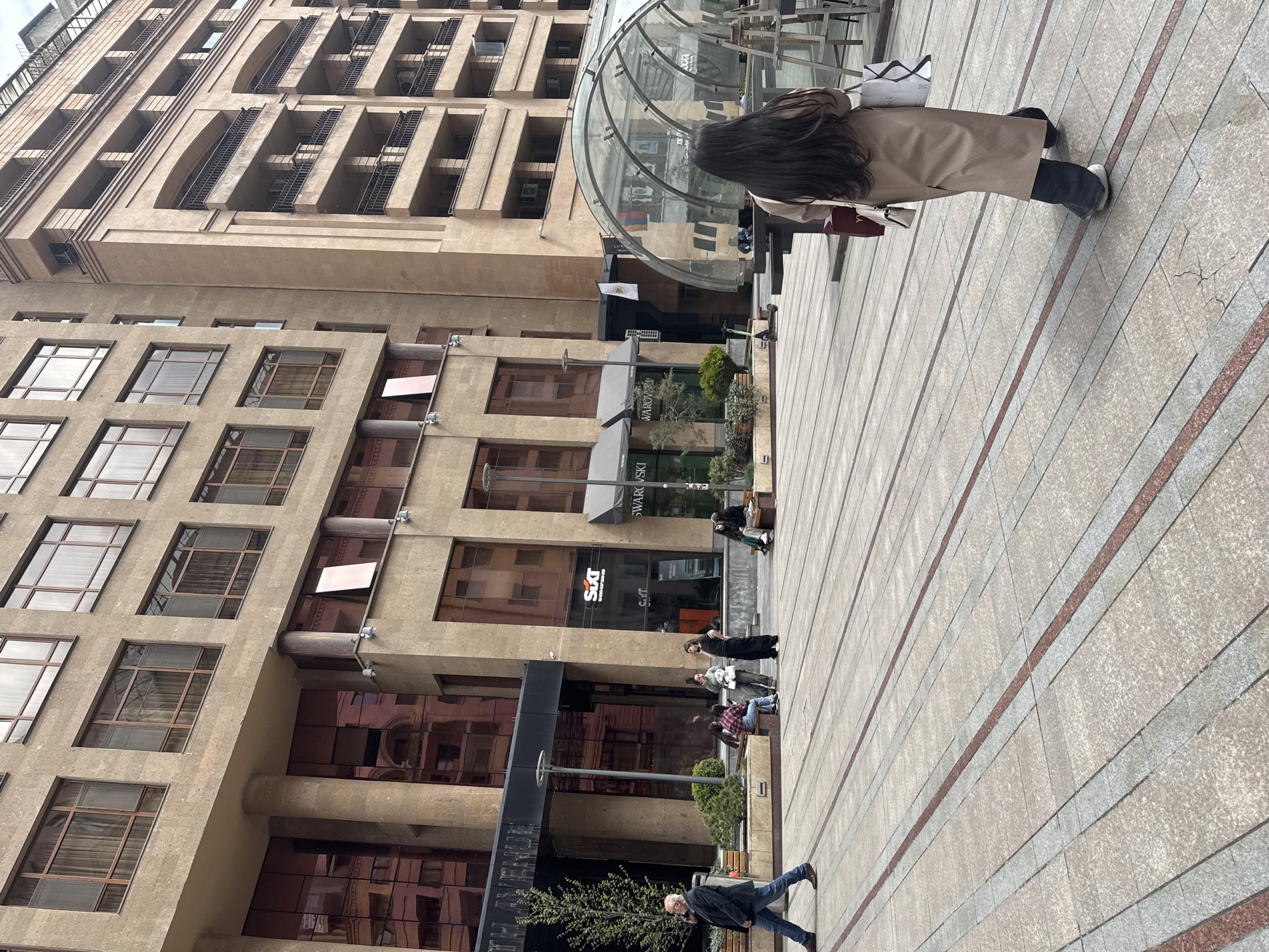The image depicts a street scene in Yerevan, Armenia. A tall, beige building with numerous windows dominates the left side of the frame. The building's architecture features decorative columns and projecting balconies. On the ground level, the building houses a "Swarovski" store and "Sixt" rental car office. Pedestrians are scattered along the paved plaza, including a woman wearing a trench coat walking away from the camera, people seated on benches, and a man walking in the lower right corner. A glass-covered structure, likely an entrance to an underground area, is visible in the upper center. The ground is paved with light-colored tiles with red accents. The overall perspective is slightly tilted.