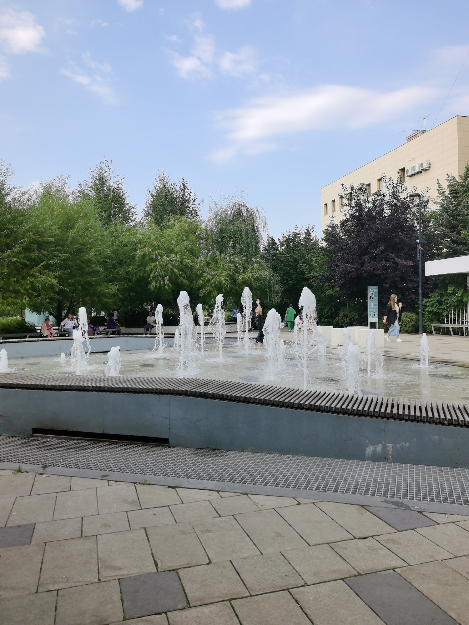 The image depicts an outdoor fountain area in a city, likely Kazan, Russia given the provided context. The fountain is the central feature, with multiple jets of water spraying upwards from a shallow pool. The pool is surrounded by a light blue concrete wall topped with a wooden slatted edge. A metal grate runs along the base of the wall. The foreground shows a paved area made of large square tiles, some of which are a darker gray. In the background, there are lush green trees and a modern, light-colored building with several windows and air conditioning units visible. Several people are scattered around the scene. Some are seated on benches in the shade of the trees, while others are walking in the vicinity of the fountain. Two women are clearly visible walking near the fountain, one holding a drink and the other walking to the right of the frame. A person in a green uniform is also present. The sky above is blue with scattered white clouds. The overall atmosphere suggests a public urban space designed for relaxation and recreation.