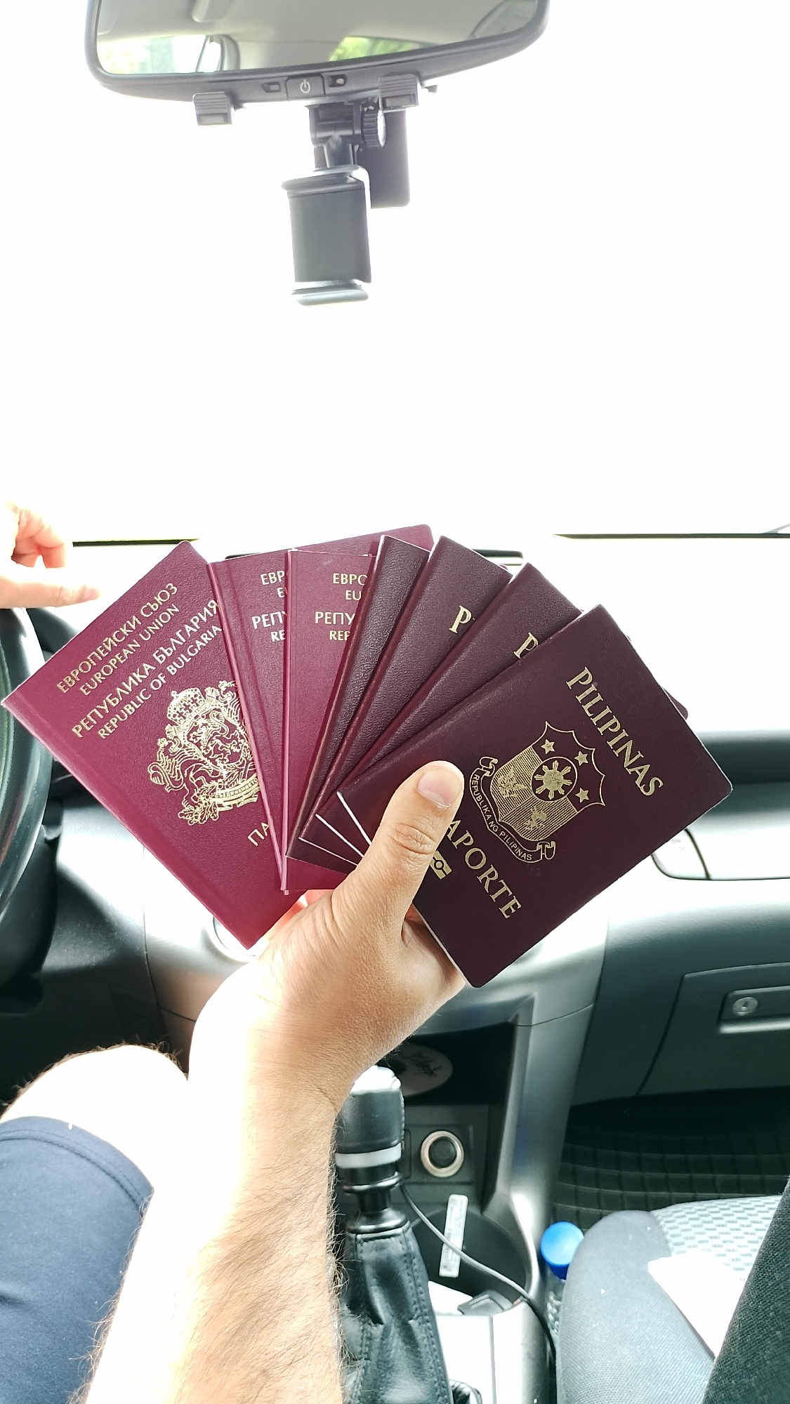In an interior shot taken from a first-person perspective inside a vehicle, a person's hand fans out multiple passports. The passports are predominantly dark red and burgundy. The foremost passport, held in the person's right hand, is a burgundy one with the word "FILIPINAS" in gold lettering across the top and the text "REPUBLIKA NG PILIPINAS" below the Philippine coat of arms. To the left of this, a fanned group of dark red passports are visible, with some featuring text in Cyrillic and English that appears to be from the European Union and the Republic of Bulgaria. The background shows the car's interior, including a gear shift, a center console with buttons and ports, and a portion of the dashboard with a rearview mirror mounted above it. The scene is brightly lit, suggesting daylight.