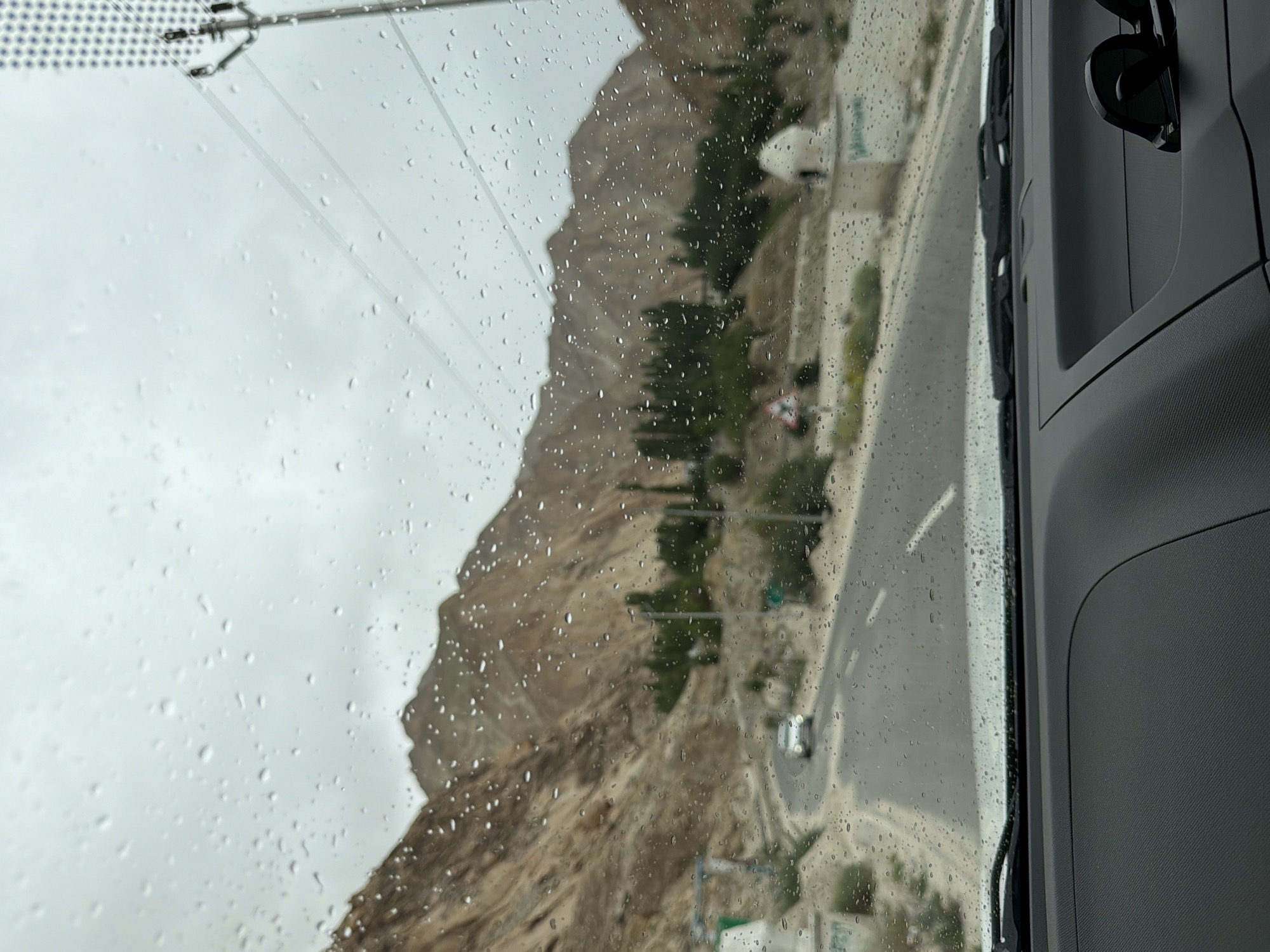 The image is taken from inside a vehicle, looking out through a rain-streaked windshield. The view is of a road winding through a mountainous, arid landscape. The sky is overcast, and the mountains are a muted brown and tan color. There are scattered green trees and some vegetation along the roadside. A single white car is visible on the road ahead, driving away from the camera. A power pole with wires is also visible on the right side of the frame. The dashboard of the vehicle is visible in the foreground, with a pair of sunglasses resting on it. The location is stated to be Ikpadok, India.