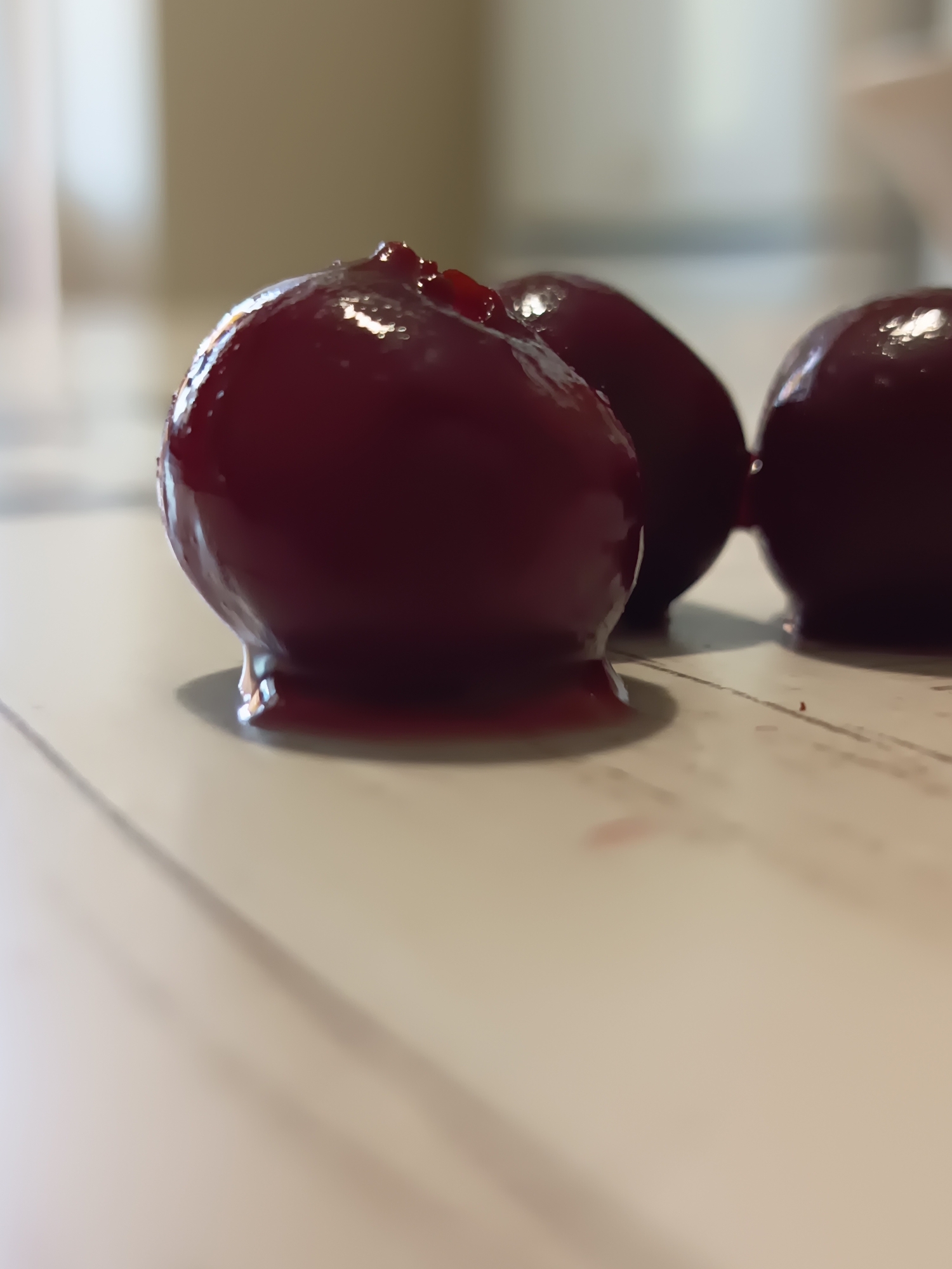 The image shows a close-up, low-angle shot of three glossy, dark red cherries placed on a light-colored, textured surface. The cherries are plump and appear to be covered in a syrup or glaze, with some of the liquid dripping down onto the surface. The lighting highlights the sheen of the cherries, creating reflections. The background is blurred, with soft, muted tones of beige and white, suggesting an indoor setting. There is no indication of the city of Izmail, Ukraine, or any specific location.