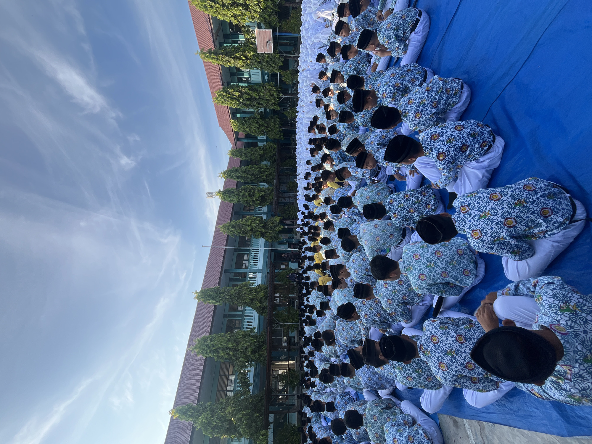 The image depicts a large gathering of male students, likely in a schoolyard setting, seated on a blue tarp. The students are predominantly wearing uniforms consisting of light-colored shirts with a repeating patterned design, white pants, and black caps. The background features a multi-story school building with red-tiled roofs and green accents, surrounded by tall, slender trees. The sky is blue with wispy white clouds. The scene appears to be an outdoor event, possibly a school assembly or ceremony, taking place during daylight hours. The perspective is from behind and slightly above the seated students, looking towards the school building. The location is identified as a school in the city of Lhokseumawe, Indonesia.