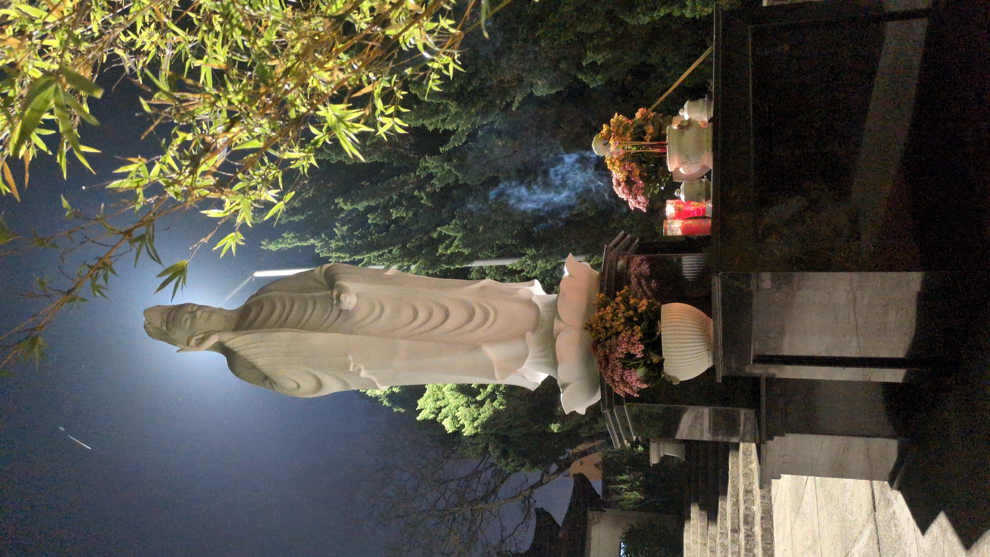 The image shows a night scene at a cemetery in Da Lat, Vietnam. A white statue of a figure, possibly a religious figure, stands prominently. In front of the statue, there are flowers in pots and on a tiered structure. Red candles are also present, with smoke visible rising from them. The tiered structure appears to be part of a tomb or memorial. The foreground has branches with leaves, likely bamboo, extending into the frame. The background consists of dark green foliage, possibly trees within the cemetery grounds. The lighting is dim, with the statue and the candles providing the main sources of illumination. The scene appears to be solemn and respectful.