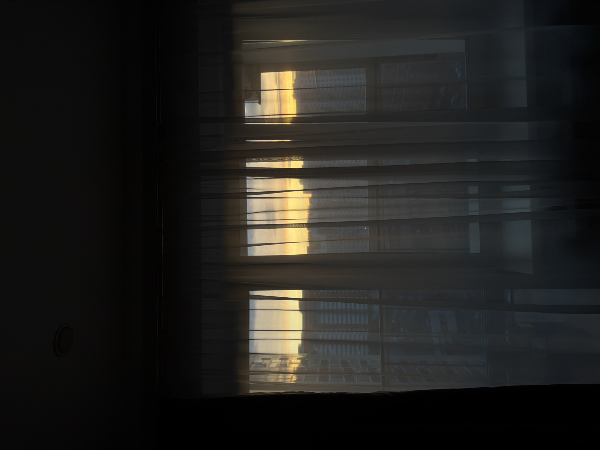 The image depicts a window in a dimly lit room. Sheer white curtains partially obscure the view outside, where a cityscape can be seen bathed in the warm glow of sunrise or sunset. The buildings visible through the window appear to be modern residential or office structures. The exact location in Krasnoyarsk, Russia is difficult to determine definitively from the image, but the architectural style and arrangement of buildings suggest a relatively new or redeveloped area of the city. The interior of the room is dark, with a portion of a wall visible to the left, featuring a simple circular element, perhaps a light switch or socket. The bottom of the window frame and a dark, undefined object are visible at the bottom of the image. There are no people visible.