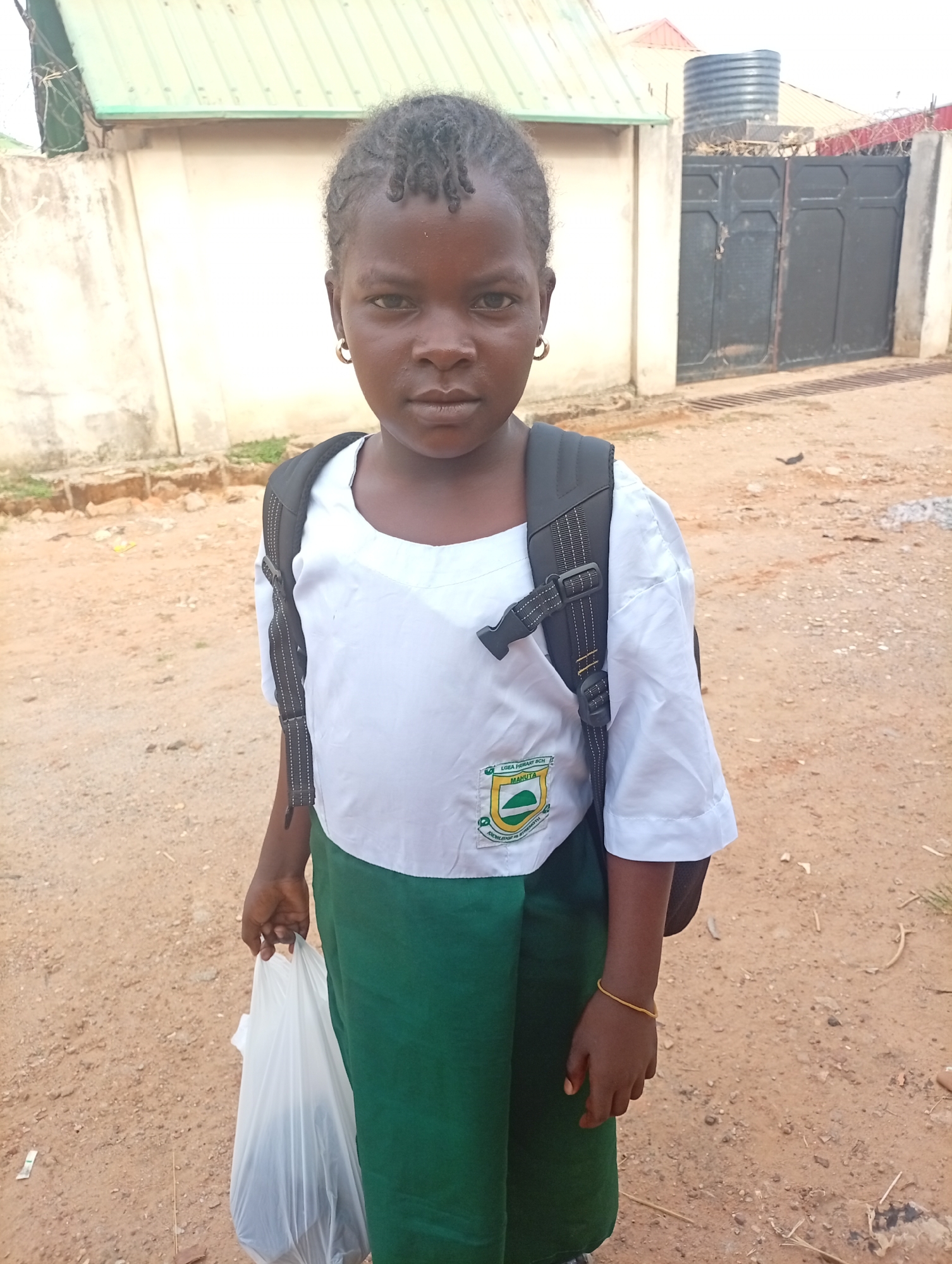 A young girl stands outdoors, wearing a white short-sleeved shirt, green skirt, and a black backpack. She is also wearing earrings and a simple bracelet. She holds a white plastic bag. The girl is in Rido, Nigeria. The background shows a building with a light green roof, a fence, and a gate. The ground appears to be dirt or gravel. A black water tank is visible on the rooftop of a nearby building.