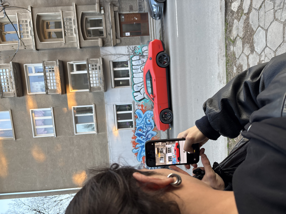 The image depicts an urban street scene in Sofia, Bulgaria, viewed from over the shoulder of an individual. In the immediate foreground, the back of an individual's head, left ear, and upper torso are visible. This person has dark hair styled back, a large silver circular ear gauge in their left earlobe, and wears a black leather jacket over a dark garment. They are holding a black smartphone vertically with both hands, positioned to photograph the street scene. The phone's screen displays a live, zoomed-out preview of the red car and the buildings behind it.
The primary subject of the smartphone's focus is a red two-door Dodge Challenger sports car, parked parallel to the curb on an asphalt street. The car features black multi-spoke wheels. Behind the Challenger, a portion of a gray vehicle, consistent with a Volkswagen Golf, is partially visible.
The background consists of two multi-story residential buildings. The building on the left is a light-brown stucco structure with several white-framed windows; its lower section is covered extensively with blue and white Cyrillic graffiti. The adjacent building on the right is a darker stucco, featuring more elaborate architectural details including ornate balconies with railings and arched window frames. A dark brown wooden entrance door is visible at street level on this building. Warm, directional sunlight illuminates parts of the building facades and reflects off some windows, suggesting late afternoon or early morning. Bare tree branches are visible against the sky on the far left. The sidewalk in the mid-ground is paved with irregularly shaped light-colored stones.