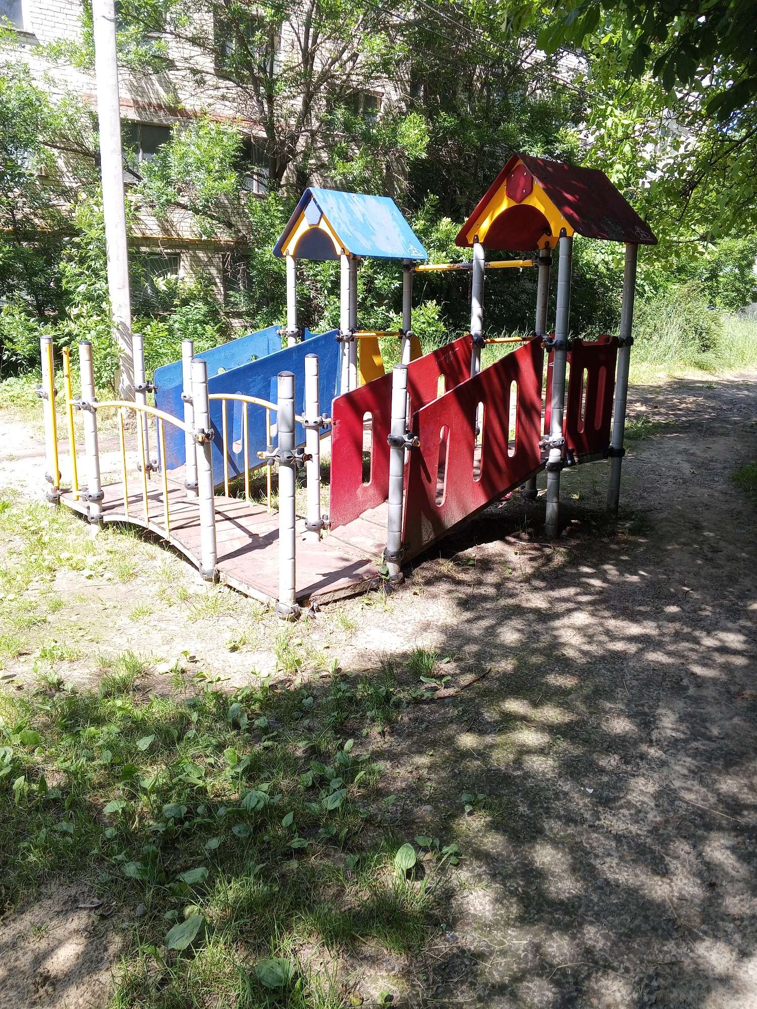 An outdoor shot captures a children's playground, featuring a colorful play structure. The structure includes a red-roofed section with yellow trim and a blue-roofed section. Connecting these is a wooden bridge with yellow railings. Red panels form a barrier on one side of the structure. The playground is situated amidst greenery, with trees and grass surrounding it. In the background, a brick building is visible, partially obscured by foliage. The ground is a mixture of dirt and grass, with dappled sunlight filtering through the trees, casting shadows on the scene. This playground is located in Novomar'evskaya, Russia. There are no people in the image.