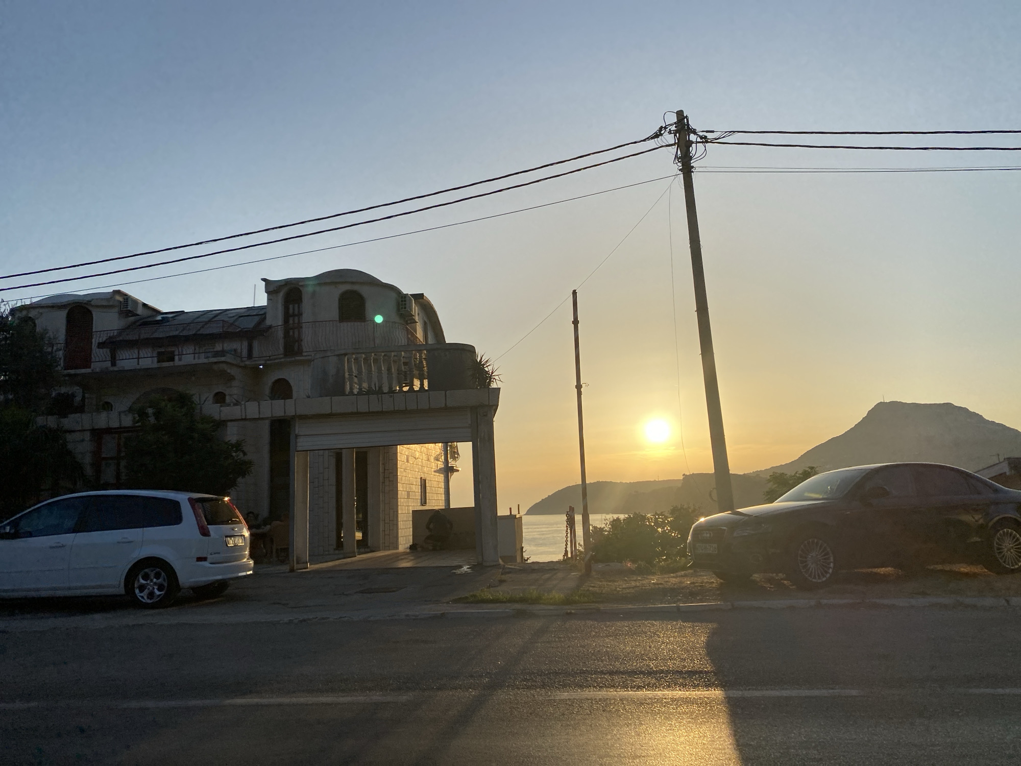 A wide-angle, eye-level, outdoor shot captures a sunset scene in Sutomore, Montenegro, featuring a modern building and two cars parked by the roadside. The sky is a clear, pale blue, transitioning to a warm yellow hue near the horizon where the sun is setting. The sun itself is a bright, small, yellow orb, casting a golden glow over the landscape. In the mid-ground, a contemporary building with curved architectural elements, possibly a residence or a hotel, is visible on the left. It has balconies and arched windows, with a partially open garage-like entrance. A white minivan is parked in front of this building, facing left. On the right side of the frame, a dark-colored sedan is parked, facing right. The road in the foreground is paved, with distinct lane markings visible. Beyond the road, a body of water, likely the Adriatic Sea, is visible, with a distant mountainous coastline silhouetted against the sunset. A utility pole with power lines runs across the upper portion of the image. The overall atmosphere is serene and picturesque, highlighting a typical coastal evening in the region.