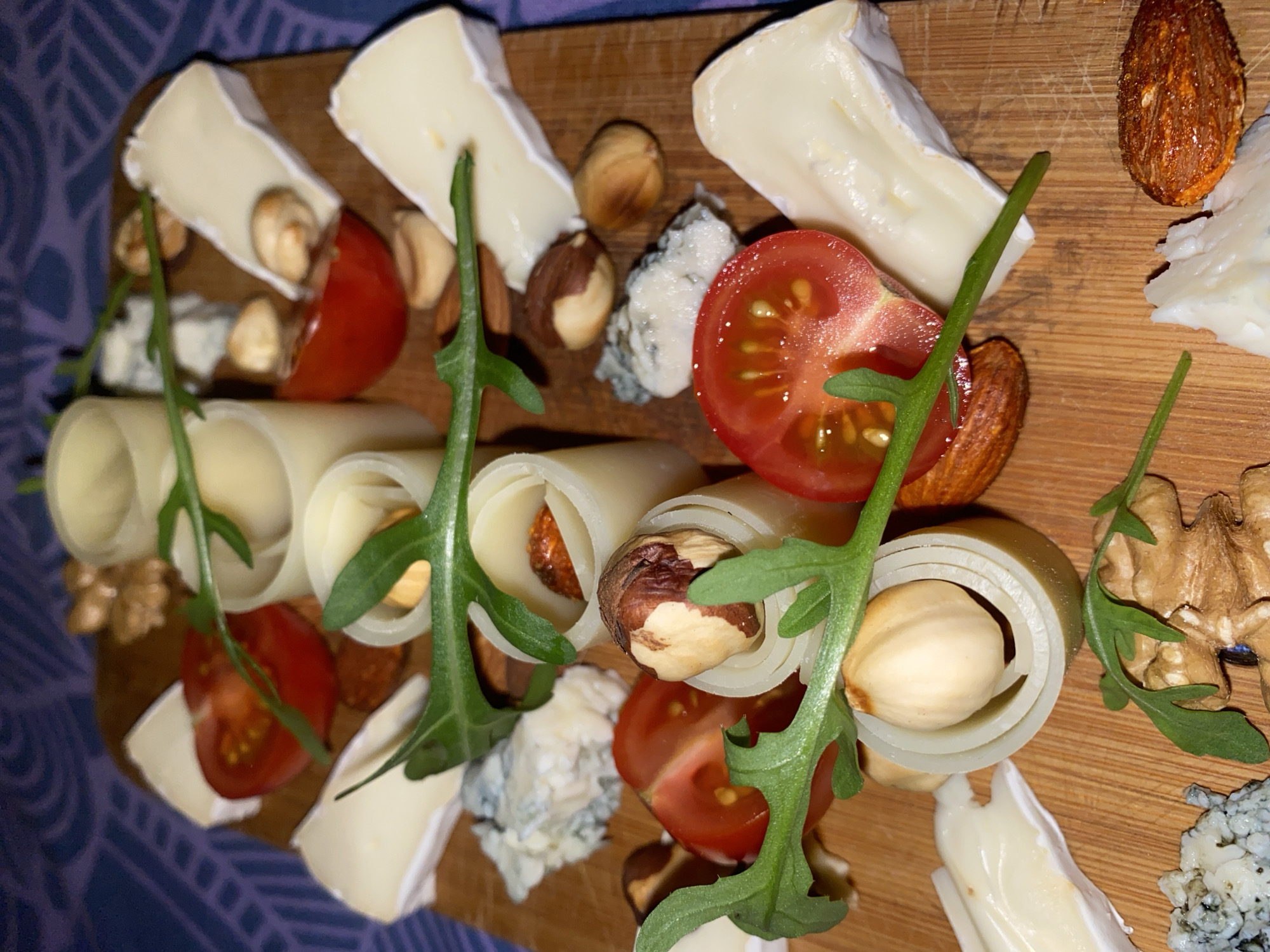 A top-down, close-up shot depicts a wooden cheese board with a variety of cheeses, nuts, tomatoes, and green sprigs, possibly arugula. Types of cheeses include soft cheeses with a white rind, blue cheese, and rolled cheese slices with a nut inside each. Nuts include hazelnuts, walnuts, and almonds. Cut cherry tomatoes add color. Green sprigs are scattered around the board. The board rests on a surface with a purple and white patterned design. The location is not specified. There is no indication that the image was taken in Chaikovsky, Russia. No people are visible in the image.