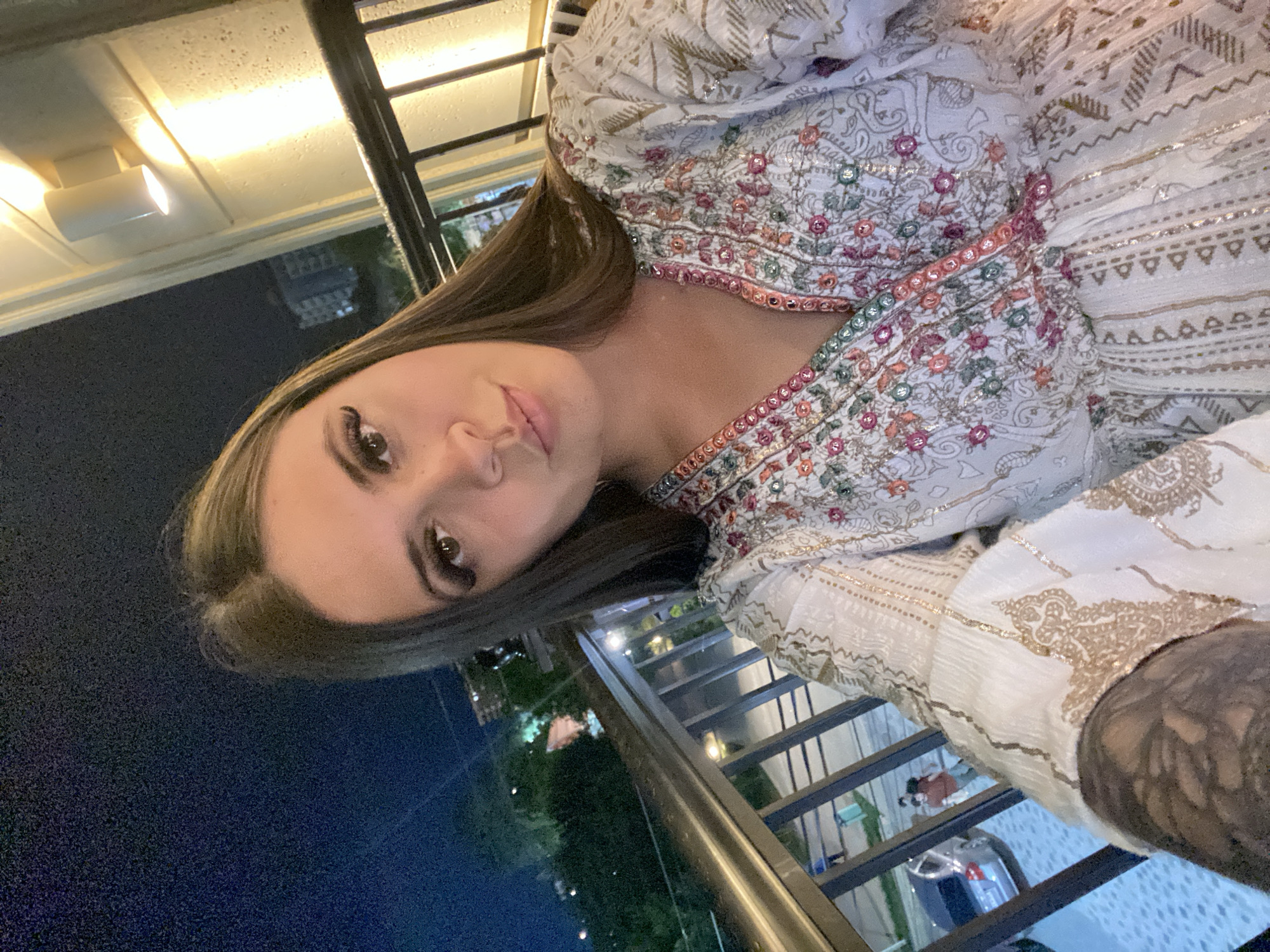An outdoor nighttime selfie is taken on a balcony in Derbent, Russia. A young woman with long brown hair and dramatic false eyelashes is the primary subject. She is wearing a white dress adorned with colorful embroidered floral patterns and metallic accents. The background shows a railing, a building with exterior lighting, and a dimly lit urban landscape with trees and distant structures. A sliver of a car and two individuals are visible on a lower level, suggesting a public space. The lighting suggests artificial illumination from lamps and ambient city lights.