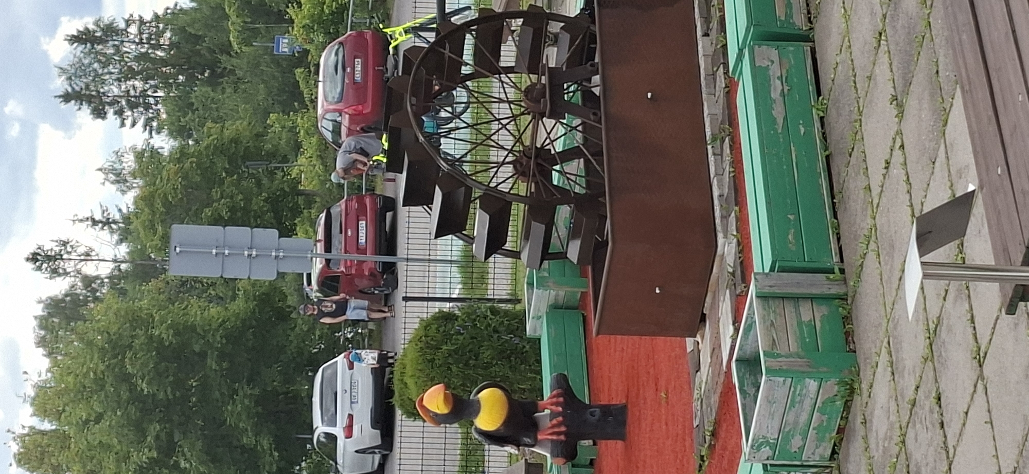 In Lahti, Finland, an outdoor mini-golf course features a decorative black bird statue with orange and yellow accents, standing on a tree trunk. Beyond it, a large, rusty water wheel mechanism sits in a rectangular trough. The course has red artificial turf with scattered green planter boxes. In the background, several cars are parked, including a white sedan and two red SUVs. Two men and a child are visible near the cars. A street sign and lush trees complete the scene.