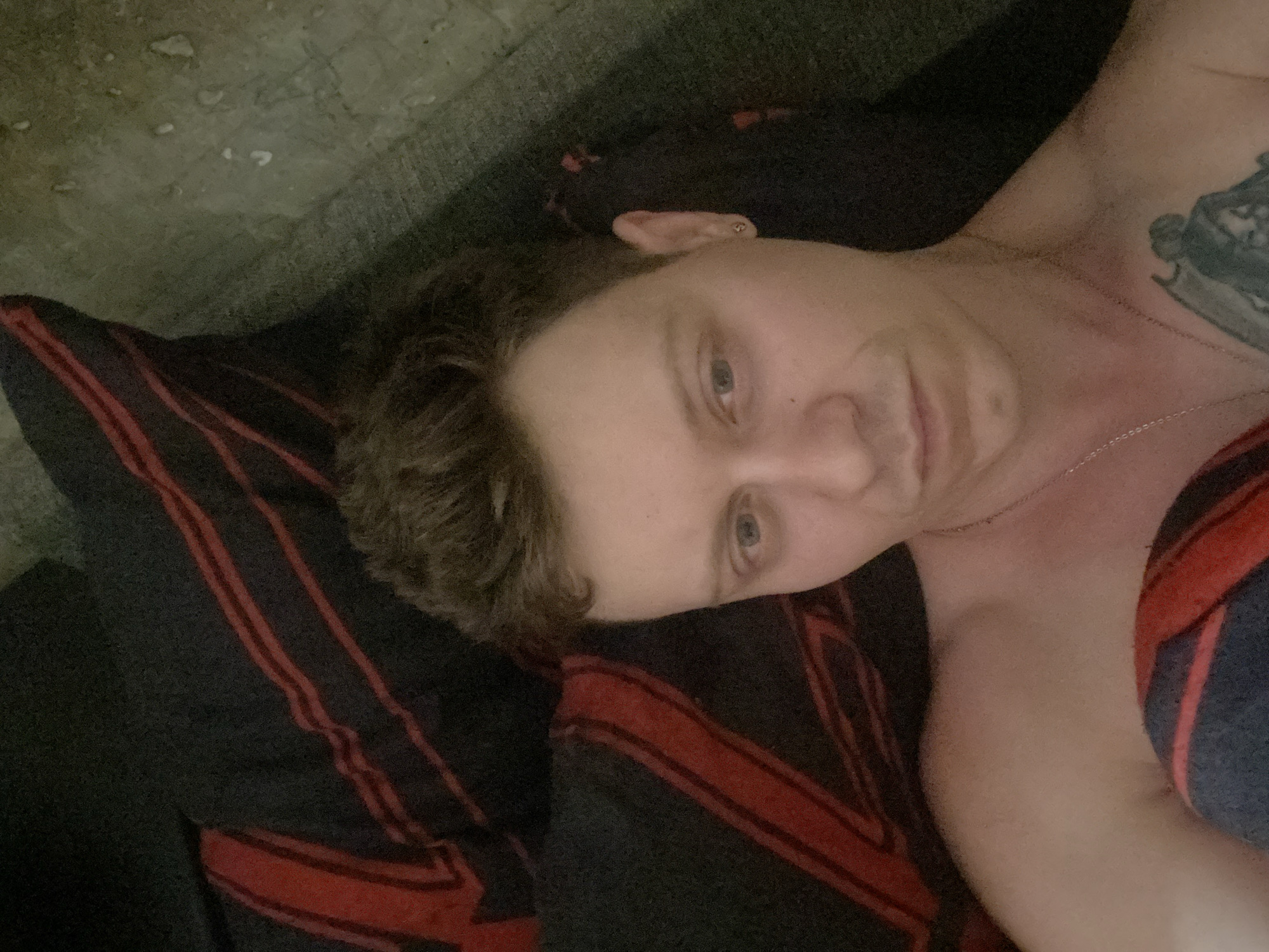 The image depicts a close-up, high-angle shot of a fair-skinned individual lying down. The person has short, dark brown hair, a light complexion, and light blue eyes. They are wearing a silver chain necklace and a small silver stud earring in their right ear. A tattoo is partially visible on their left shoulder, appearing to depict a winged figure. The background consists of dark fabric with red stripes, possibly a blanket or bedding, and a textured, light-colored surface that could be a wall or floor. The lighting is dim, creating shadows that obscure some details. The image does not provide any identifiable landmarks or contextual information that would allow for the determination of its location in the city of Samara, Russia.