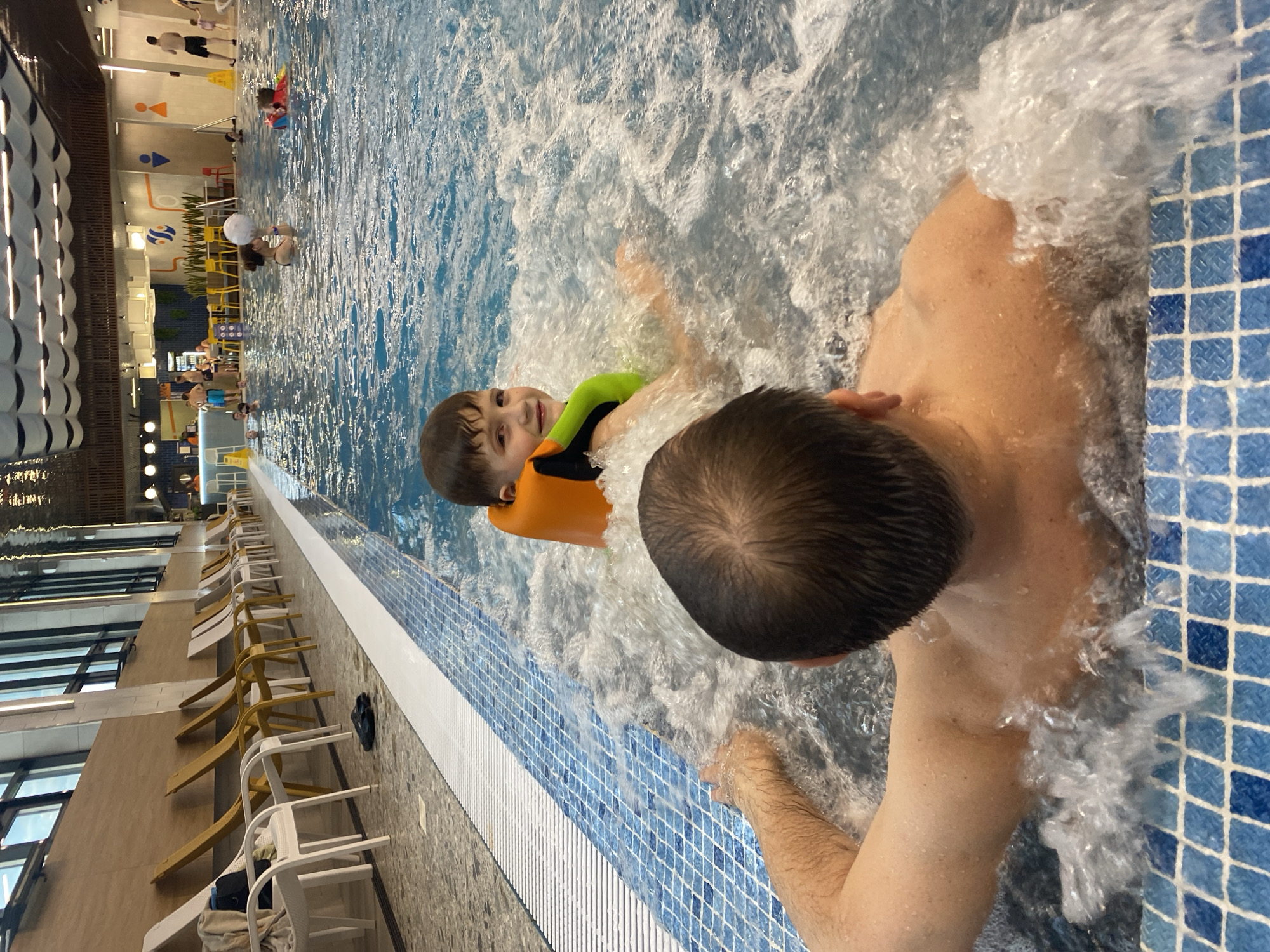 This image depicts an indoor swimming pool facility. In the foreground, an adult's head and shoulders are visible, facing away from the camera, with their arm reaching into the bubbly water of a hot tub or jacuzzi. To the left, a young boy wearing a bright orange and green life vest is seated in the bubbly water, looking towards the camera with a smile. He appears to be in the same hot tub as the adult. The pool area features blue and white checkered tiling along the edge. In the background, there is a row of lounge chairs and other swimmers in a larger pool. The facility has high ceilings with modern lighting fixtures. The scene is set within the city of Votkinsk, Russia.