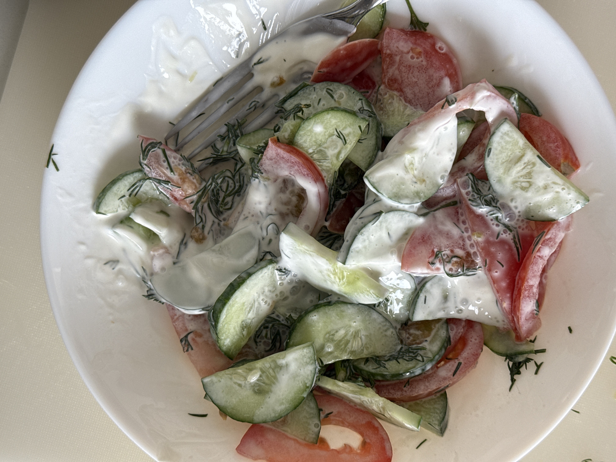 A top-down shot shows a white bowl filled with a cucumber and tomato salad dressed in a creamy, white sauce. The salad includes sliced cucumbers and tomatoes, with dill sprigs scattered throughout. A silver fork is placed on top of the salad. The location in the city of Dikaya Gar', Russia, is not evident from the image.