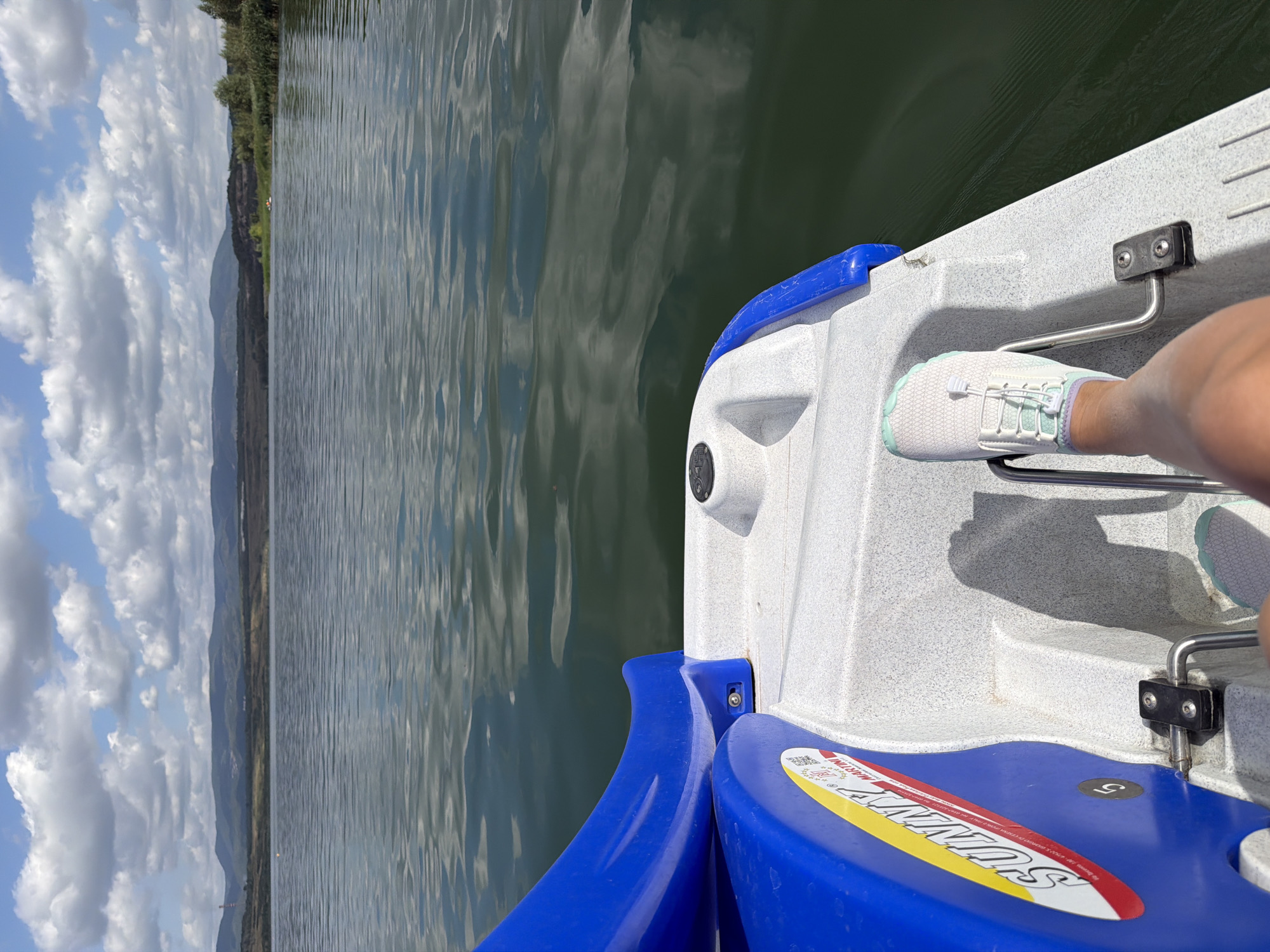 The image shows a first-person perspective of someone on a paddleboat on a lake. The paddleboat is white and blue, with a "SUNNY" logo visible on the side. One foot, wearing a white sneaker with light blue and purple accents, is on a pedal. The lake's surface is rippled and reflects the sky, which is partly cloudy. In the background, there is a shoreline with trees and hills, and a vast, hazy mountain range under a bright blue sky with scattered white clouds. The overall impression is a leisurely activity on a calm body of water with a scenic backdrop. The location is likely one of the reservoirs near the city of Most, Czechia, such as the Most Reservoir or Lake Matylda, which are popular recreational areas offering paddleboat rentals and surrounded by hills.
