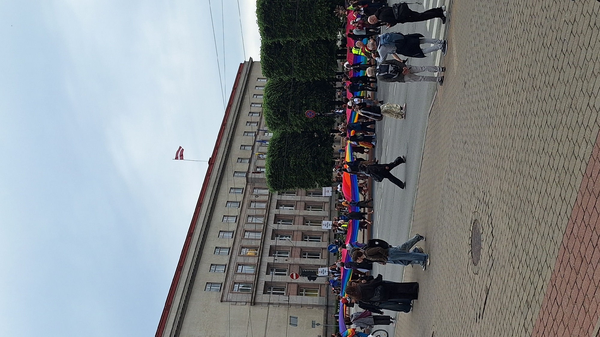 The image depicts a street scene in Riga, Latvia, likely during a Pride event. A large rainbow flag is being held by a group of people walking across a street in front of a substantial, light-colored building. The building appears to be an institutional or governmental structure, with numerous windows and a red-tiled roof. A Latvian flag is visible flying from a flagpole on top of the building. Several trees with dense green foliage are planted in a line between the building and the street. A crowd of people are walking in the street, some carrying the rainbow flag, while others are standing on the sidewalk. The street is paved with asphalt, and the sidewalk is made of brick pavers. Traffic signals and street signs are visible in the background, suggesting an urban environment. The sky is overcast with light clouds.