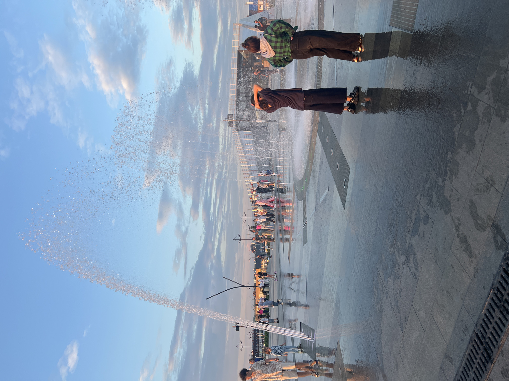 The image shows a public space with a fountain, likely in Magnitogorsk, Russia, given the context of the city being known for its public spaces and fountains. Several people are present, some interacting with the fountain's water jets. In the foreground, two individuals, one with their back to the camera wearing a dark t-shirt and wide-legged pants, and another facing away in a green plaid shirt and dark pants, stand on the wet, reflective pavement near the fountain. To the left, a young person in a light-colored shirt and shorts walks away from the fountain. Further back, a crowd of people are visible, some walking, others standing, in the background, suggesting a vibrant public gathering. The fountain itself is active, with water spraying upwards and creating a shimmering effect against the backdrop of a partly cloudy sky. The ground is wet and reflective, indicating the presence of water from the fountain. In the distance, structures and lighting characteristic of urban infrastructure are visible.