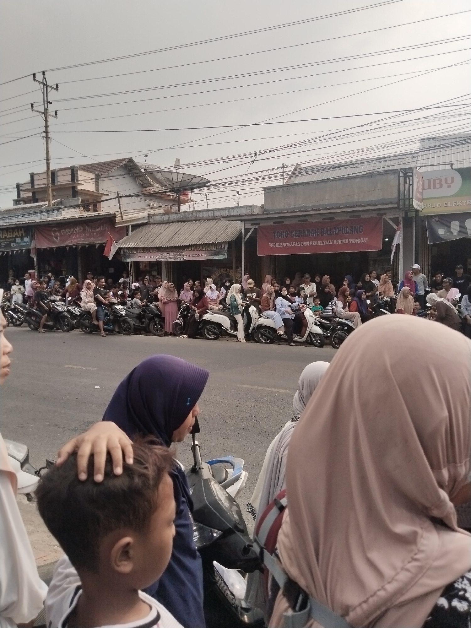 The image depicts a street scene in Balapulang Kulon, Indonesia, likely on a cloudy day. A crowd of people, many wearing headscarves, are gathered on or near motorcycles parked along the side of a paved road. Several businesses with awnings and signs line the street. One prominent red banner reads "TOKO GERABAH BALAPULANG" and "PERLENGKAPAN DAN PERALATAN RUMAH TANGGA". Another sign for "GRAND MEDIA STATIONERY" is visible. Overhead, a complex network of electrical wires crisscrosses the sky, supported by utility poles. In the foreground, the back of a child's head is visible, with a hand resting on top of it, suggesting an adult's presence. The overall impression is of a community gathering or a busy commercial area.