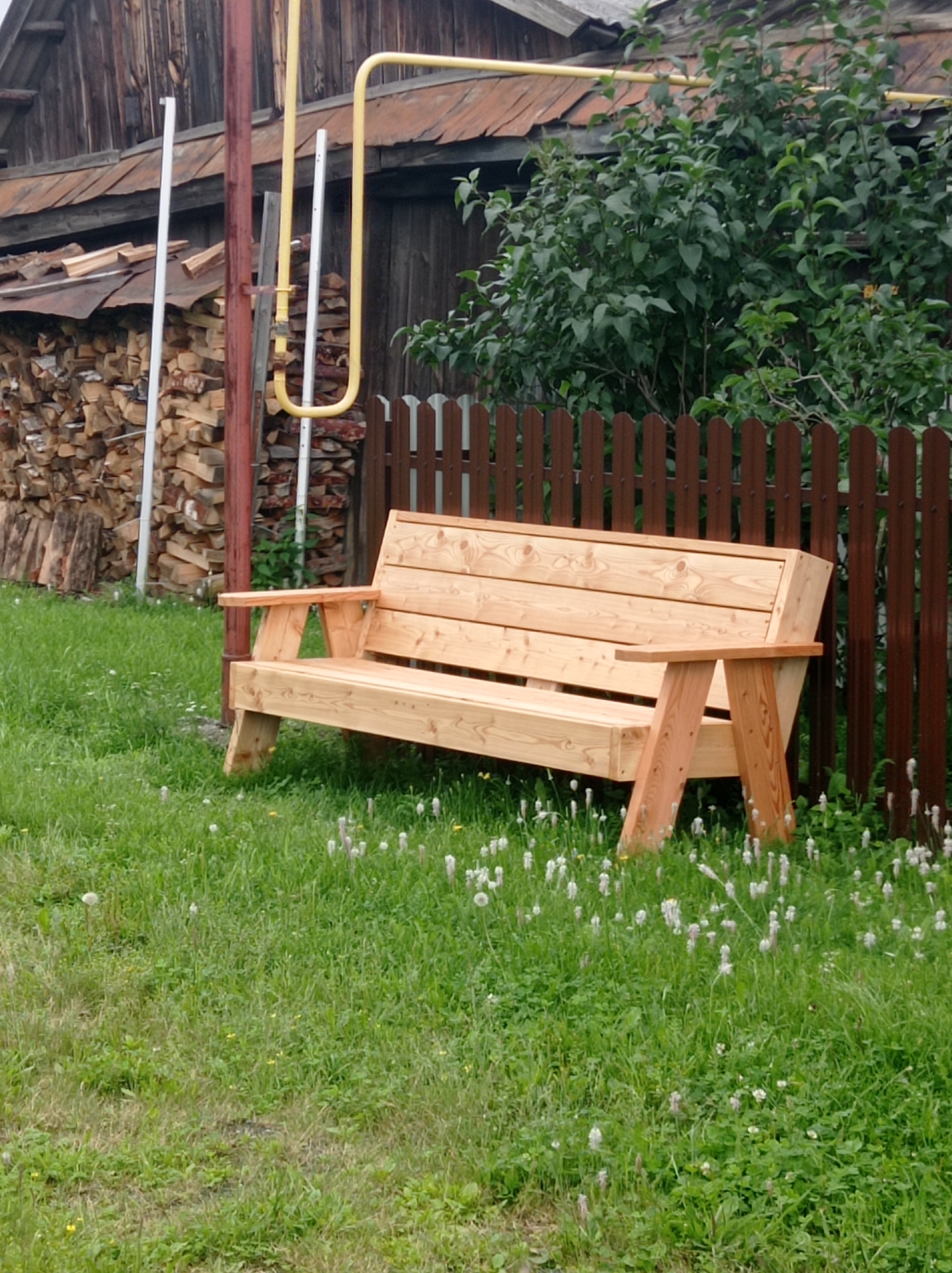 A light-colored wooden bench sits on a grassy area in front of a wooden fence and a weathered wooden building. Behind the bench, a thick green bush grows. To the left of the bench, a pile of firewood is stacked against the side of the building. A yellow pipe, likely a gas line, runs horizontally across the top of the frame, supported by vertical poles. The grass is dotted with small white flowers. The scene appears to be a rustic backyard in Degtyarsk, Russia.