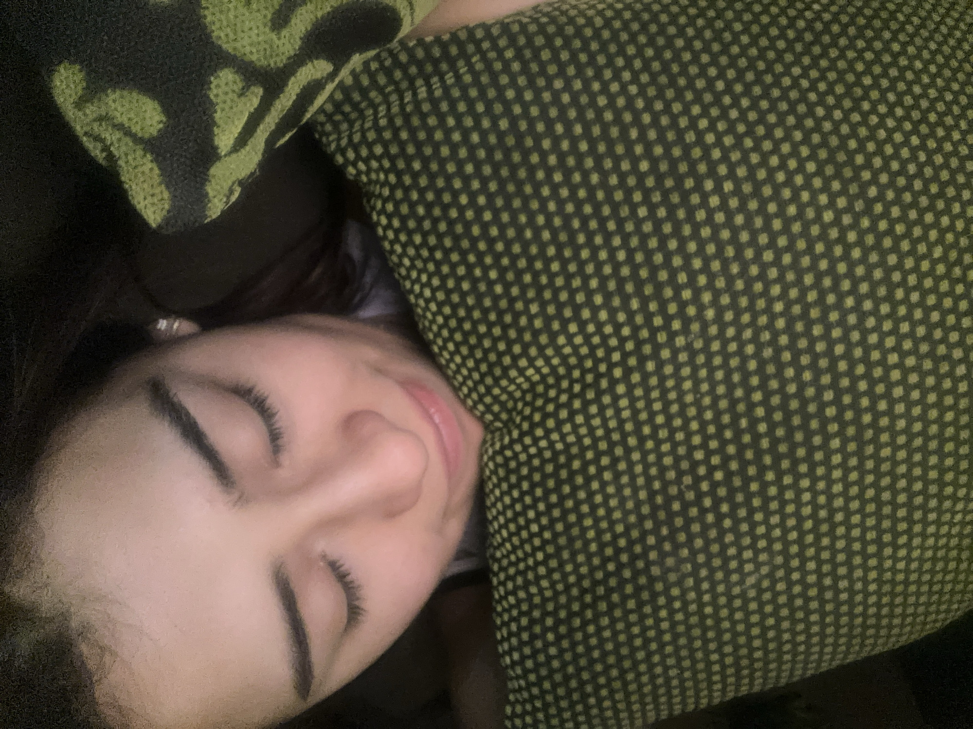 A close-up, low-angle shot shows a woman with closed eyes resting her head on a green pillow with a dark pattern. Her face is fair, with dark eyebrows and eyelashes. A pillow with a green and black pattern is visible in the upper left corner. The environment is dark. There is no evidence to determine if the location is in Puebla, Mexico.