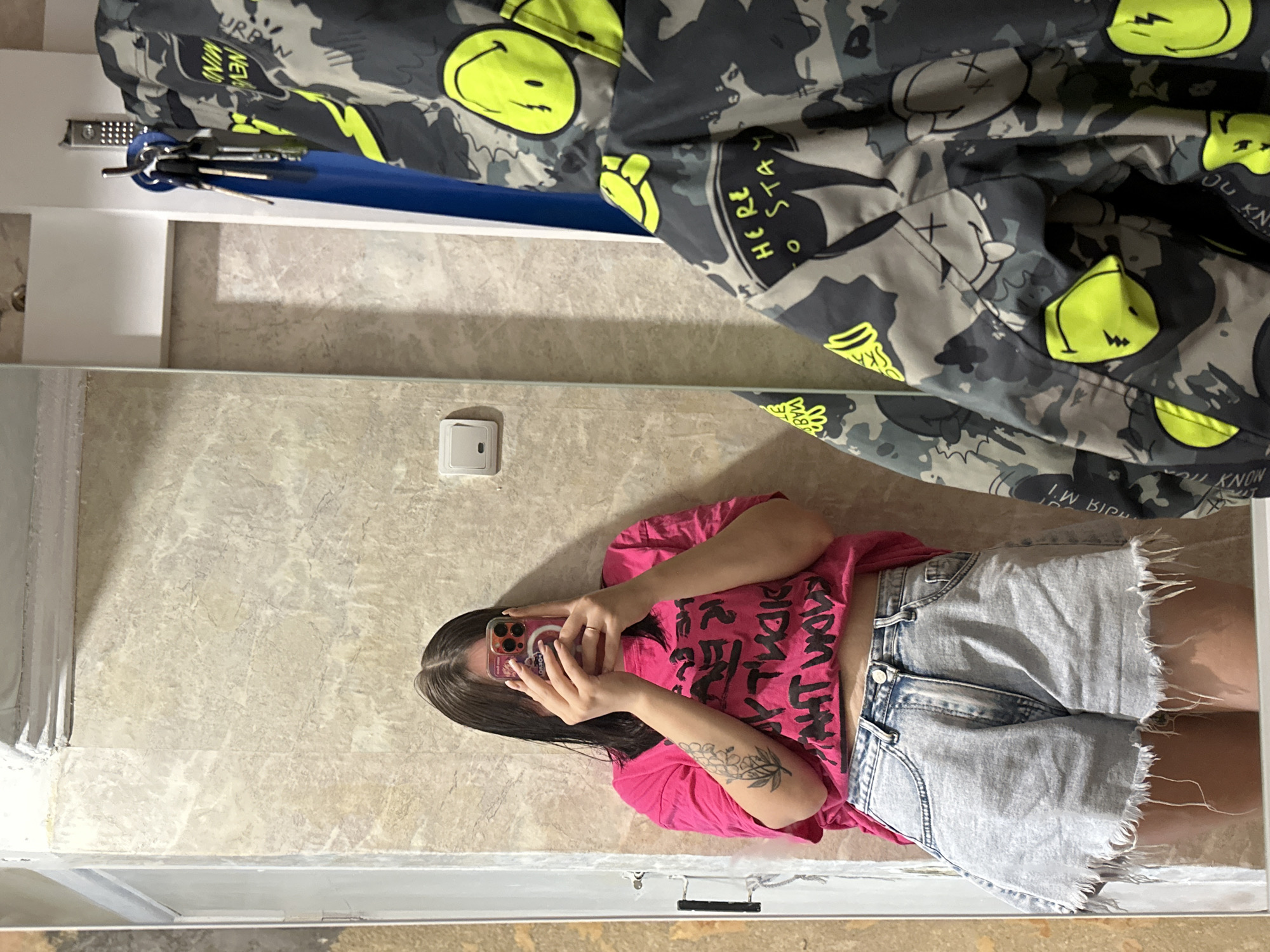 In an indoor setting, likely a bathroom or hallway with tiled walls, a person is taking a mirror selfie. The individual is wearing a bright pink t-shirt with black graphic text and light-wash denim shorts with a frayed hem. They are holding up a smartphone to obscure their face. To the right of the frame, a camouflage jacket with neon yellow smiley face and lightning bolt graphics is visible, hanging on a hook. Keys are also hanging from the hook. The overall environment appears to be domestic.